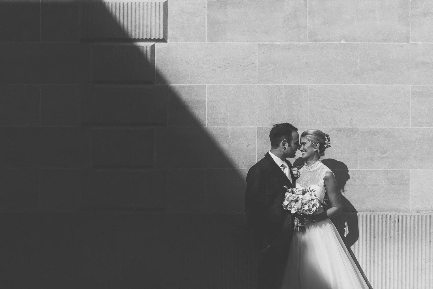 Black and white photo of a bride and groom standing close together against a wall, smiling, with their foreheads touching. The bride holds a bouquet of flowers, and sunlight creates a shadow pattern on the wall behind them.