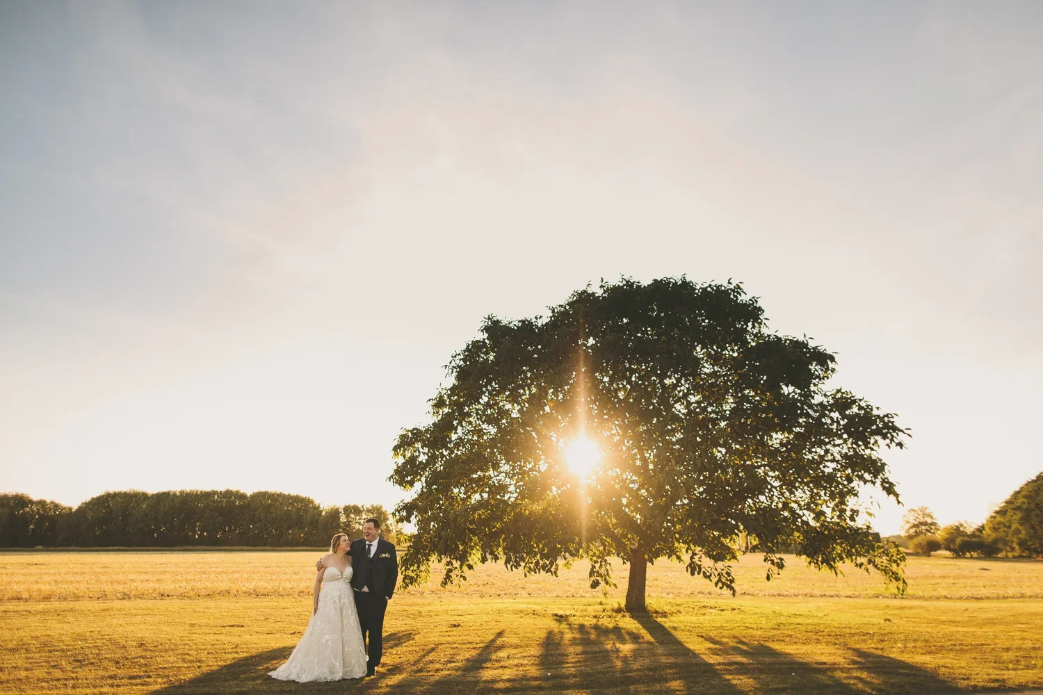 A bride and groom in wedding attire standing on grass near a large tree during sunset in a wide open field.