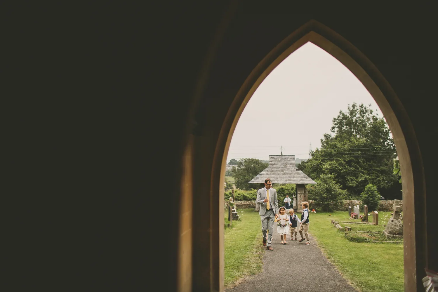 A man in a gray suit with a yellow tie walking with four children in a cemetery, seen through a church window, with trees and a small building in the background.