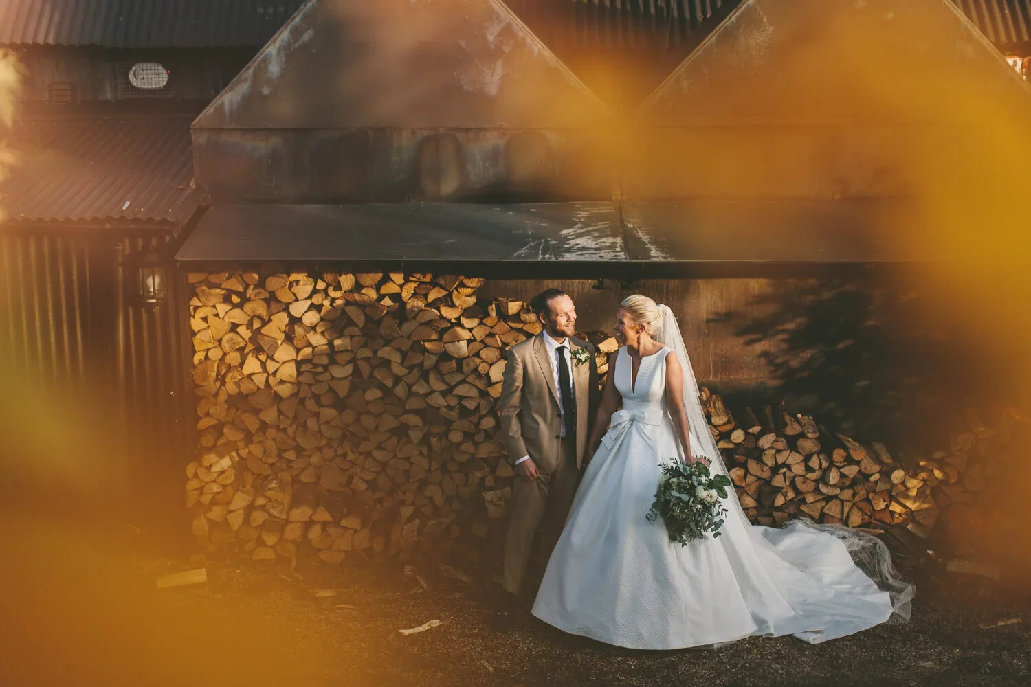 Wedding couple standing outdoors near a stack of firewood, with trees and a rustic barn in the background, as the sun sets.