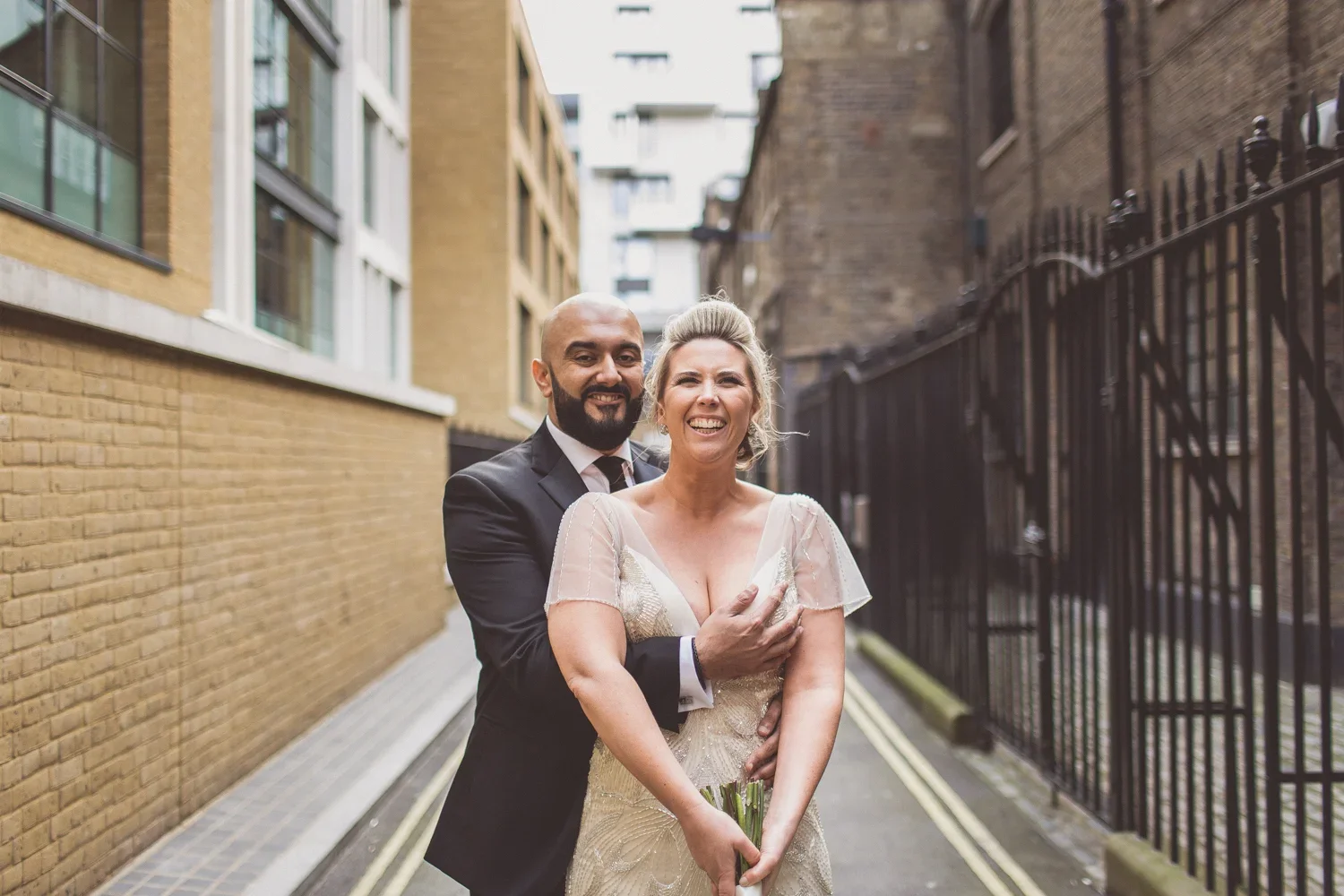 A smiling couple in wedding attire standing close together outdoors in an alleyway between brick buildings with metal fencing on one side. The woman is in a wedding dress holding a bouquet, and the man is in a black suit.