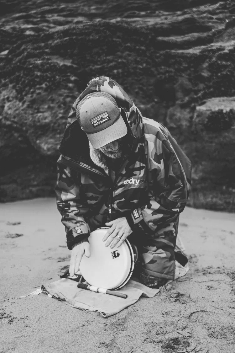 A person wearing a camouflage jacket and a Patagonia hat kneels on the sand, opening a portable cooking stove on a cloth at the beach.