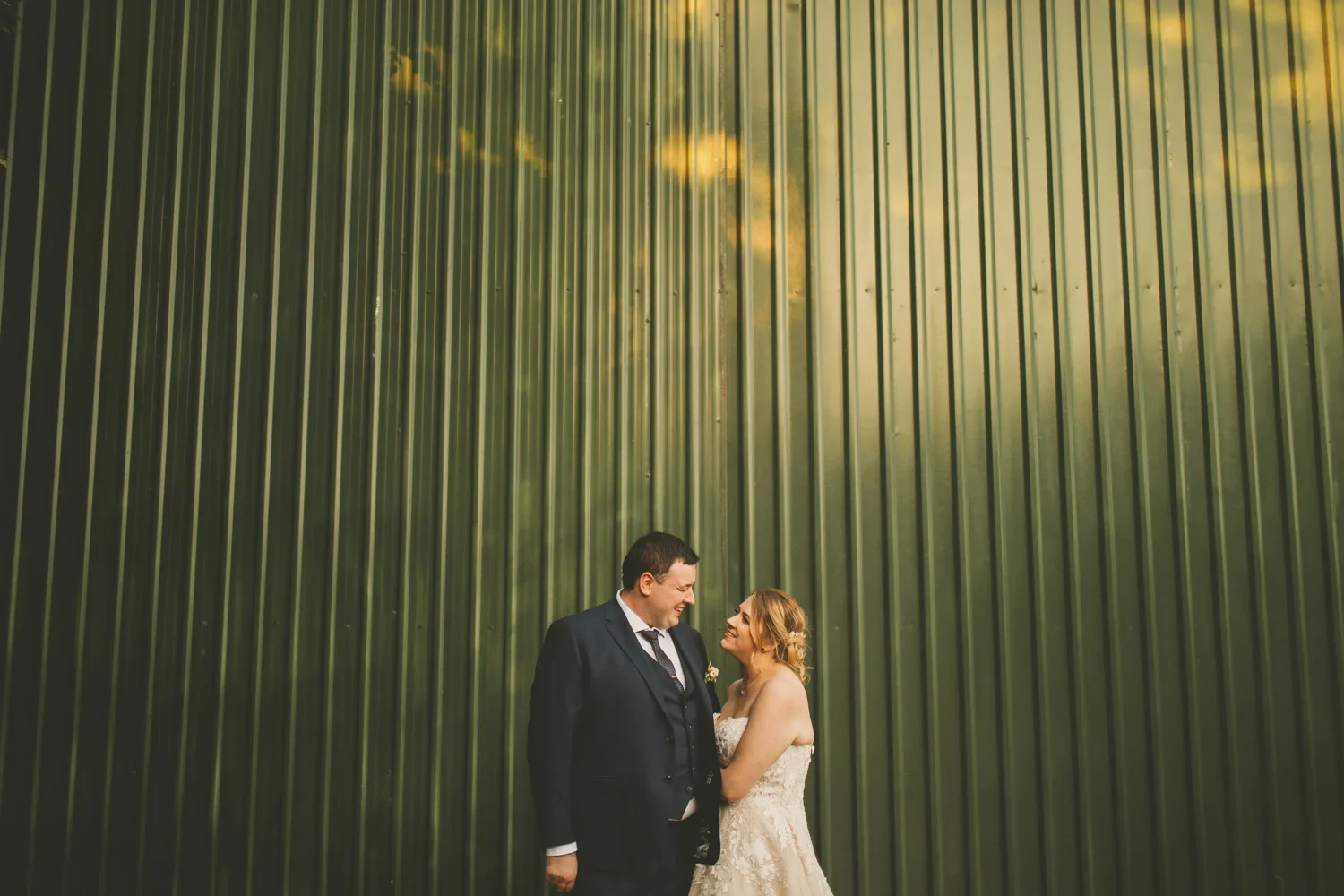 A bride and groom standing close together, smiling and looking into each other's eyes, against a large green metal wall at sunset.
