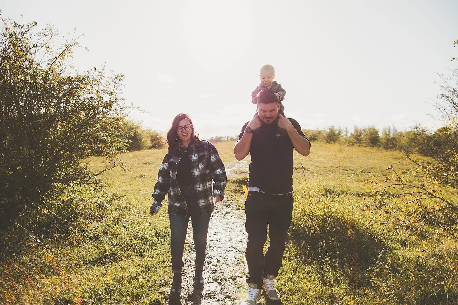 A happy family of three walking outdoors on a grassy trail during daytime, with a man carrying a young child on his shoulders and a woman smiling beside them.