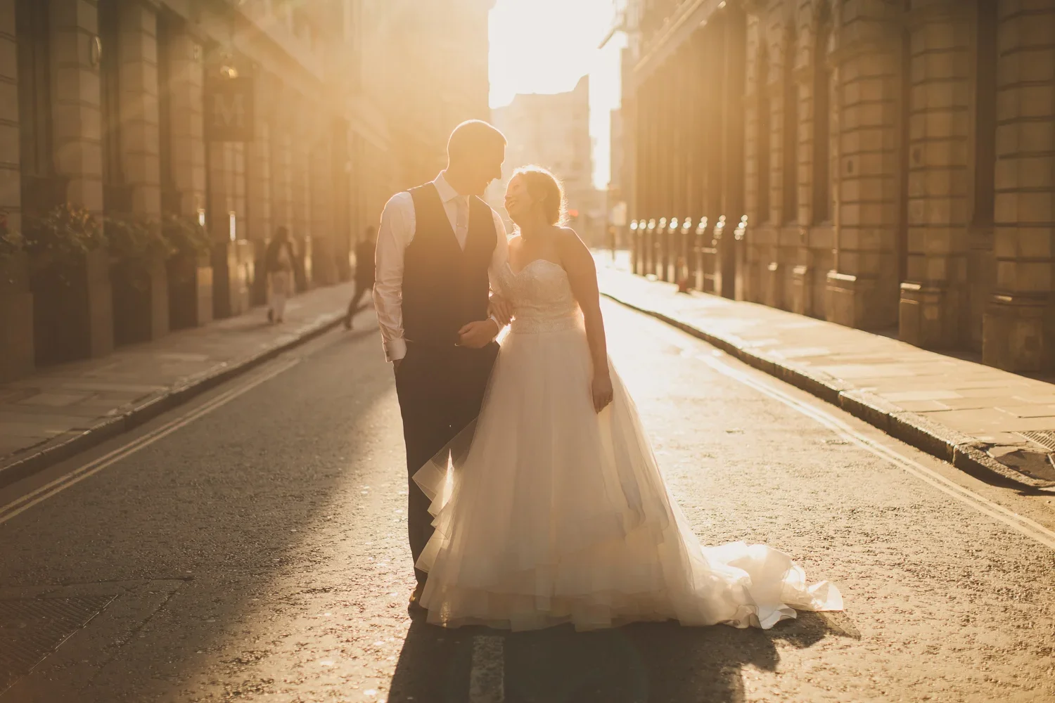 A bride and groom holding hands and smiling at each other on a city street at sunset, with the sun casting a warm glow around them.