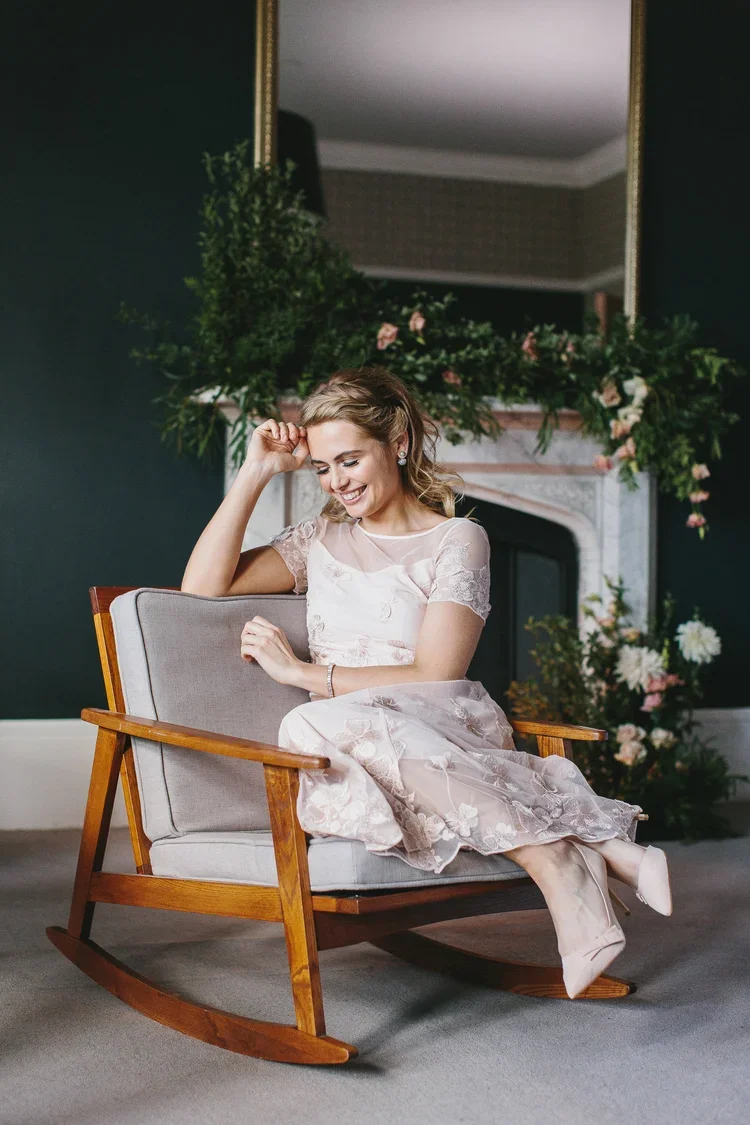 A woman in a light pink dress sitting on a wooden rocking chair, smiling, with a fireplace and floral decorations behind her.