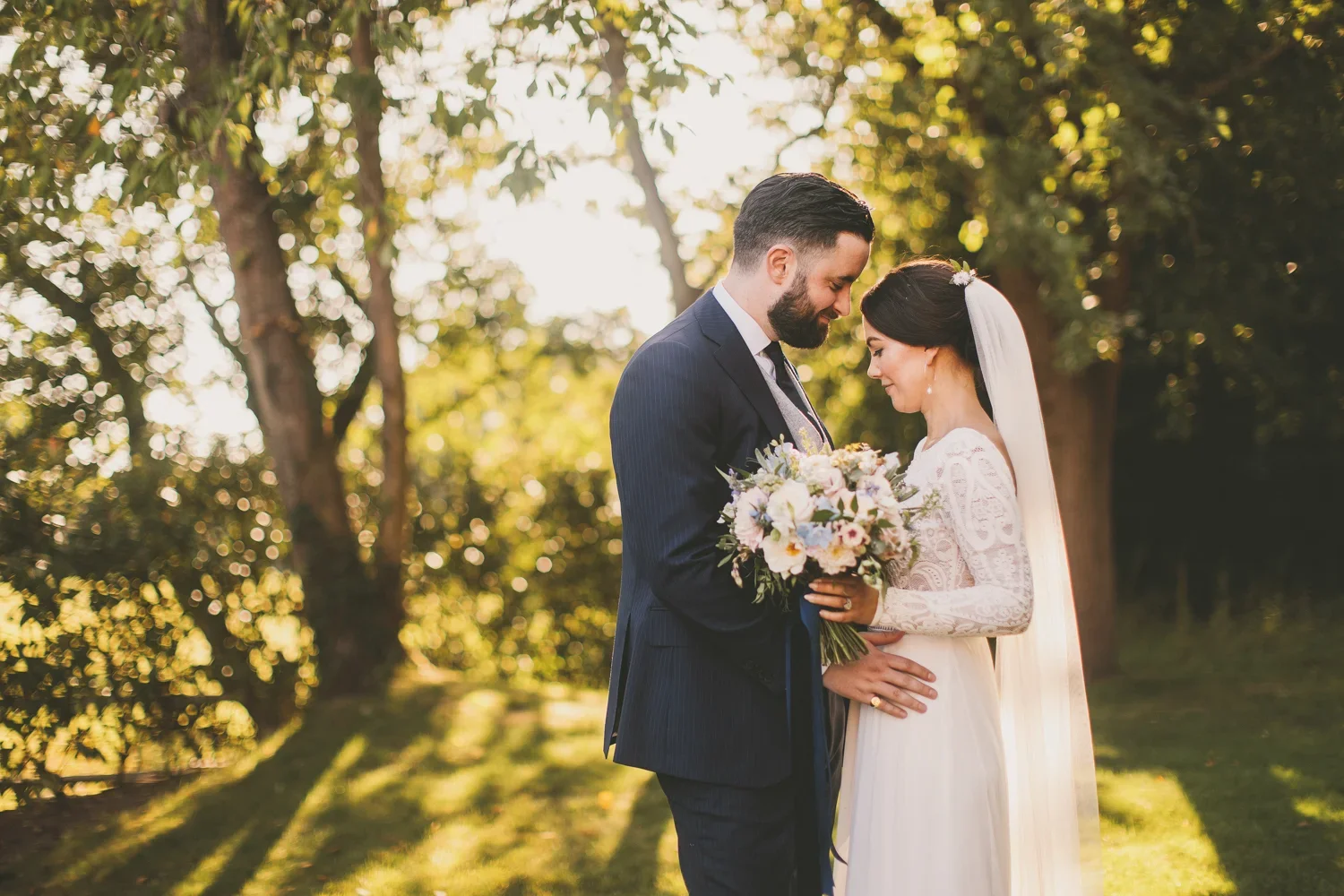 A bride and groom standing close together outdoors, holding a bouquet of flowers, during their wedding ceremony at sunset with trees in the background.