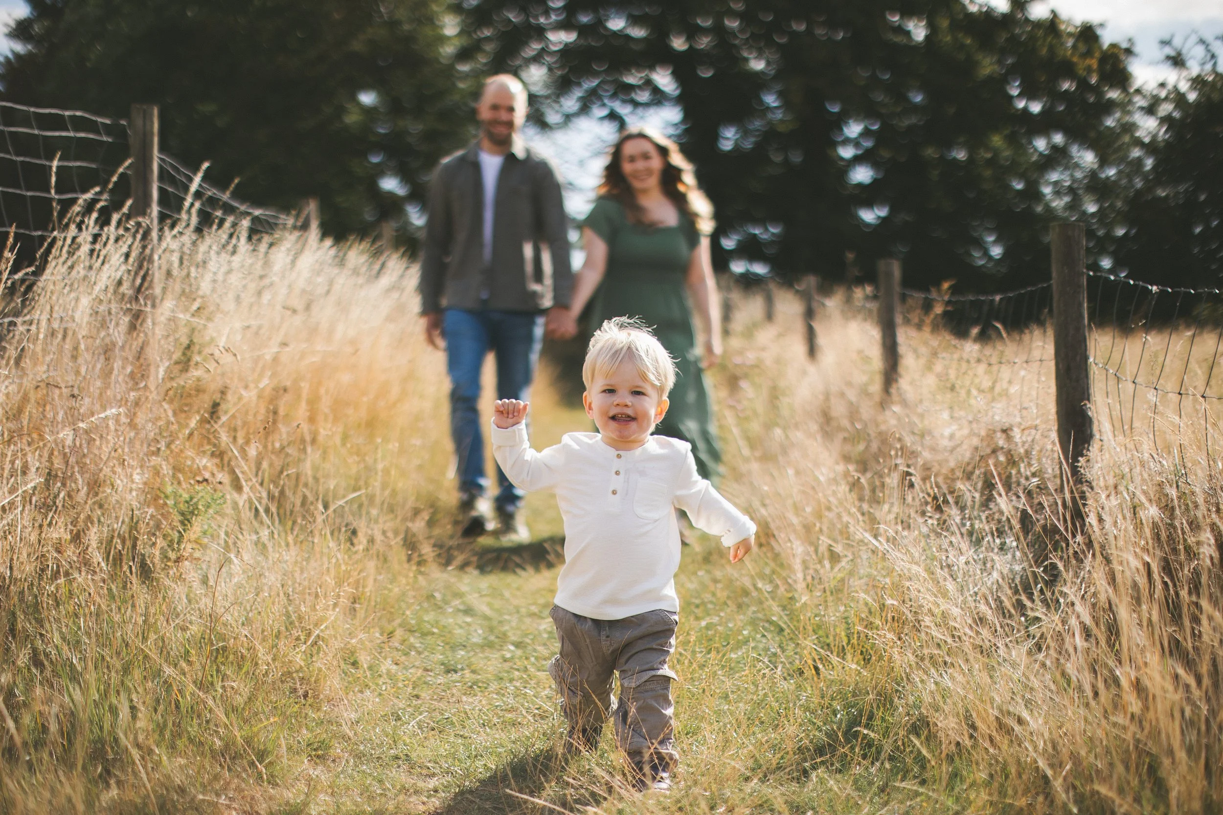 A young boy with blond hair running on a grassy trail while holding his mother and father's hands, with a woman and a man following behind him, all walking through a field with a wire fence on one side and large trees in the background.