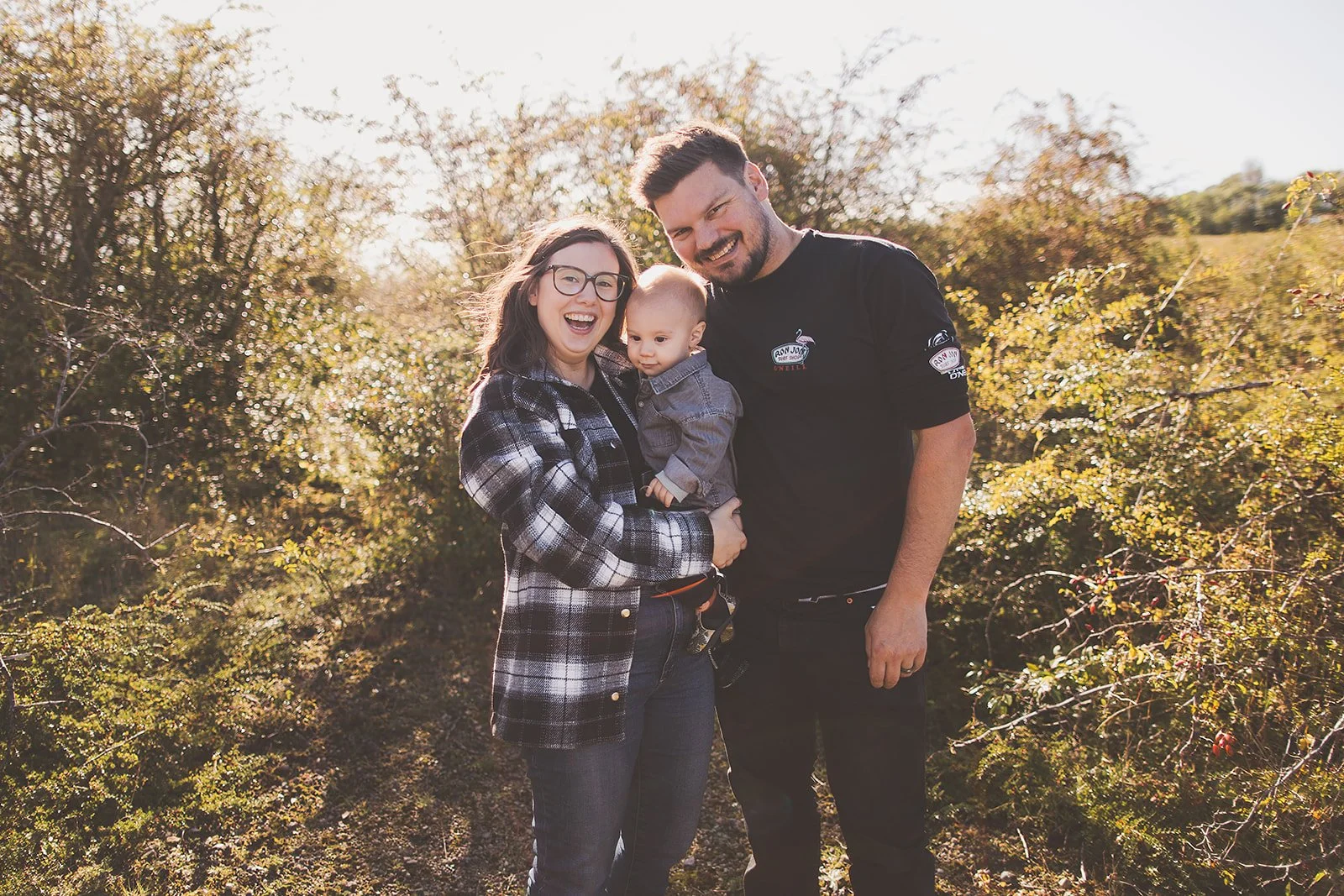 A happy family of three outdoors in autumn, smiling at the camera, with trees and sunlight in the background.