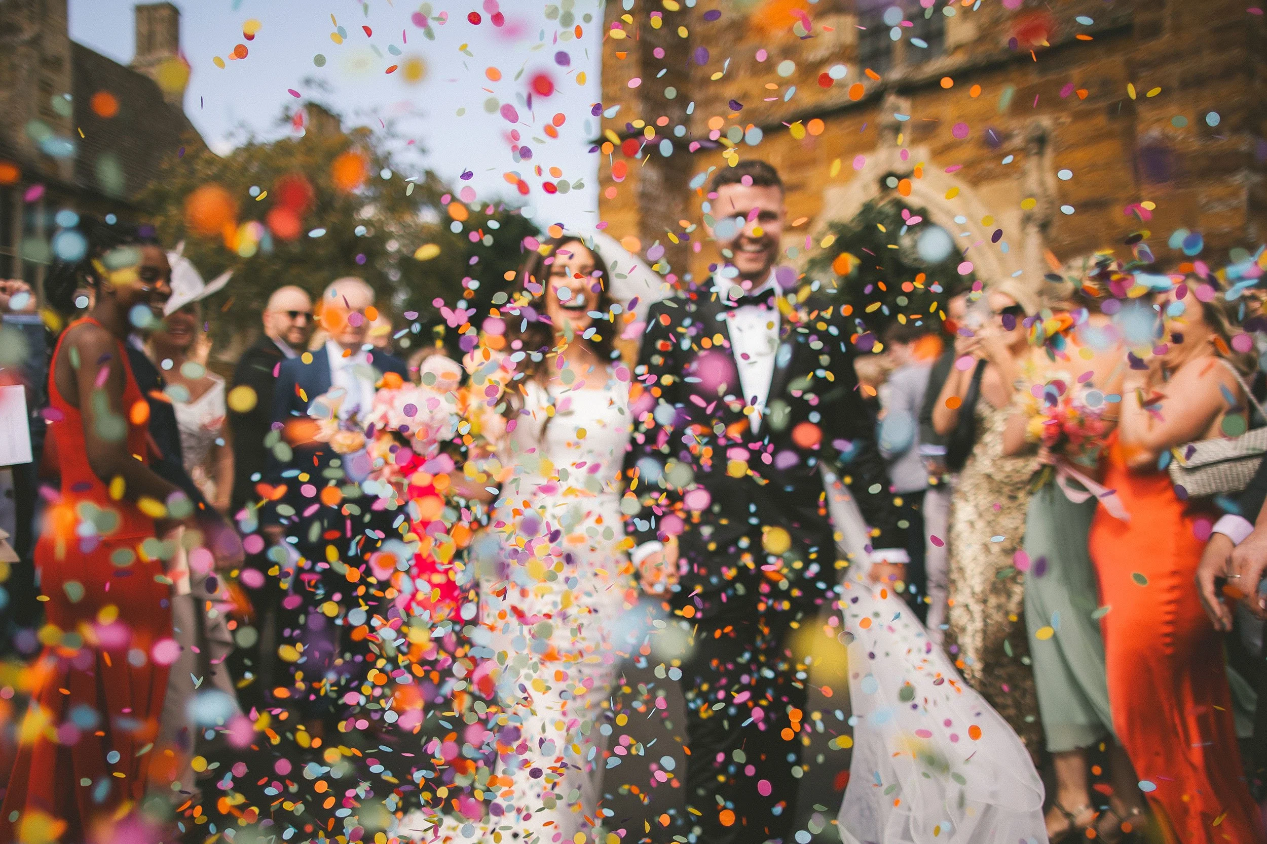 A bride and groom celebrating at their wedding reception with colorful confetti in the air, surrounded by guests.