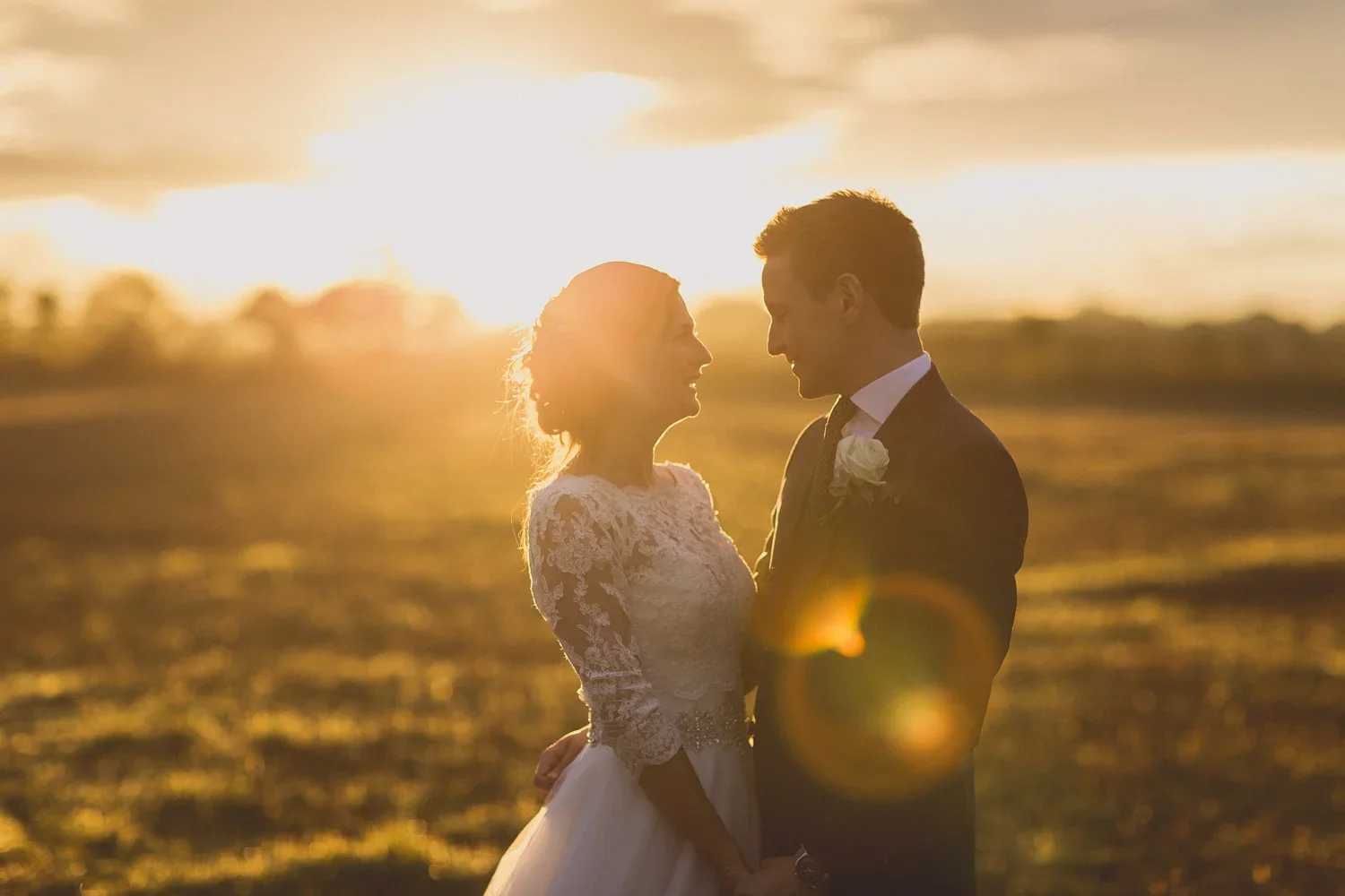 A bride and groom standing close together during sunset at a beach, smiling and gazing into each other's eyes.