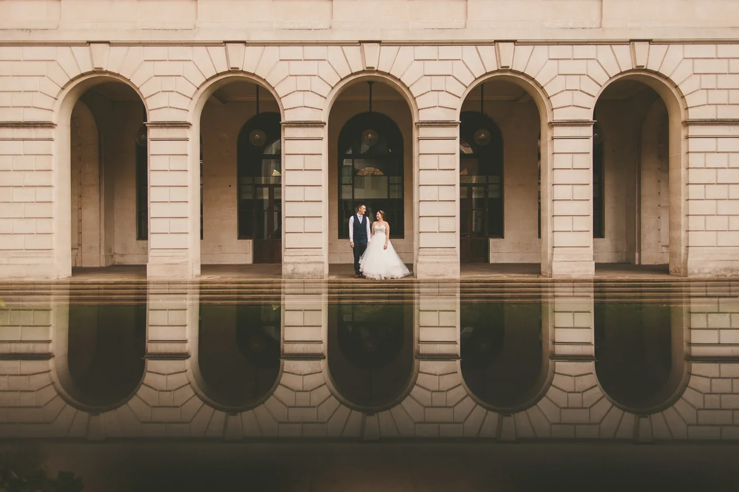 A bride and groom standing on steps in front of an elegant building with large arches, reflected in a body of water.
