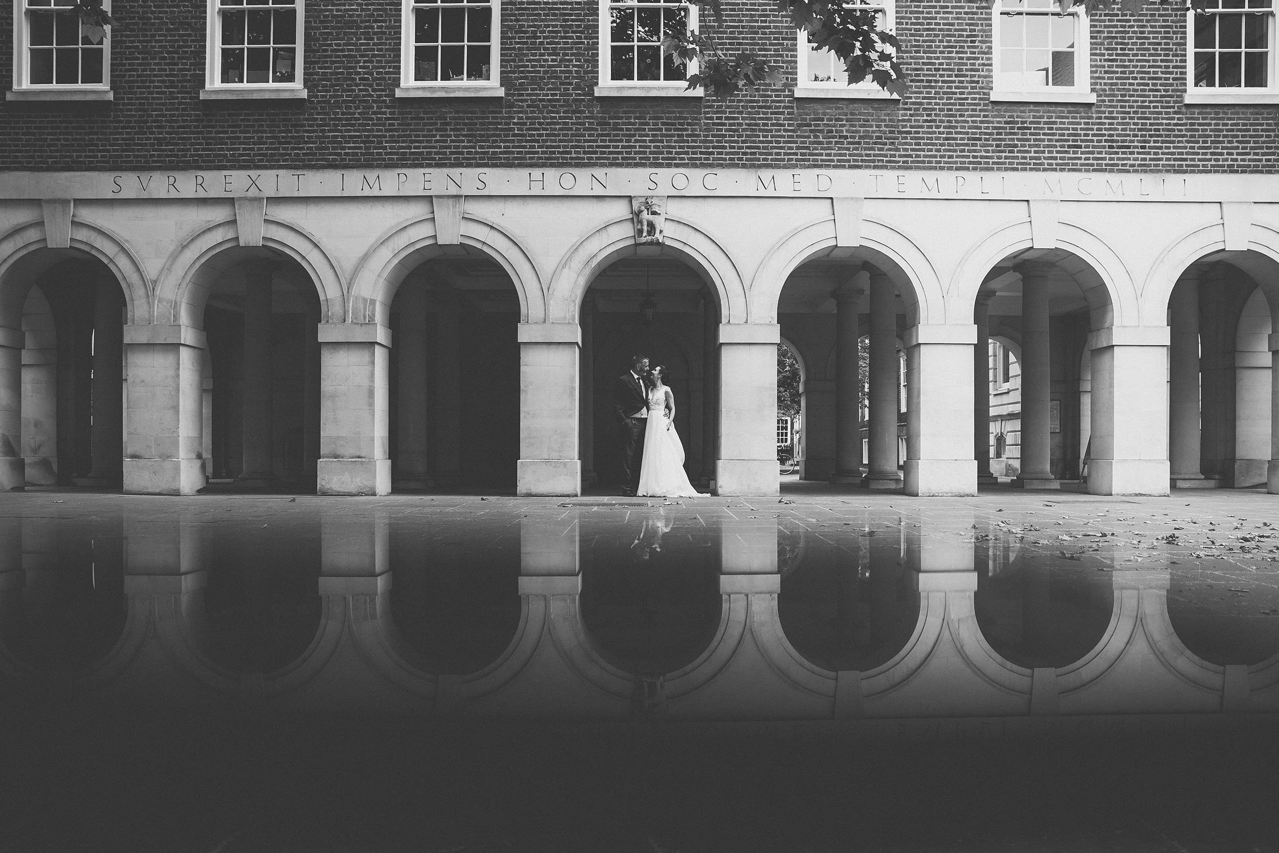 Black and white photo of a historic building with curved arches, a Latin inscription, and a statue of a lion above the arches. A couple in wedding attire stands under the arches, reflected in a body of water in the foreground.
