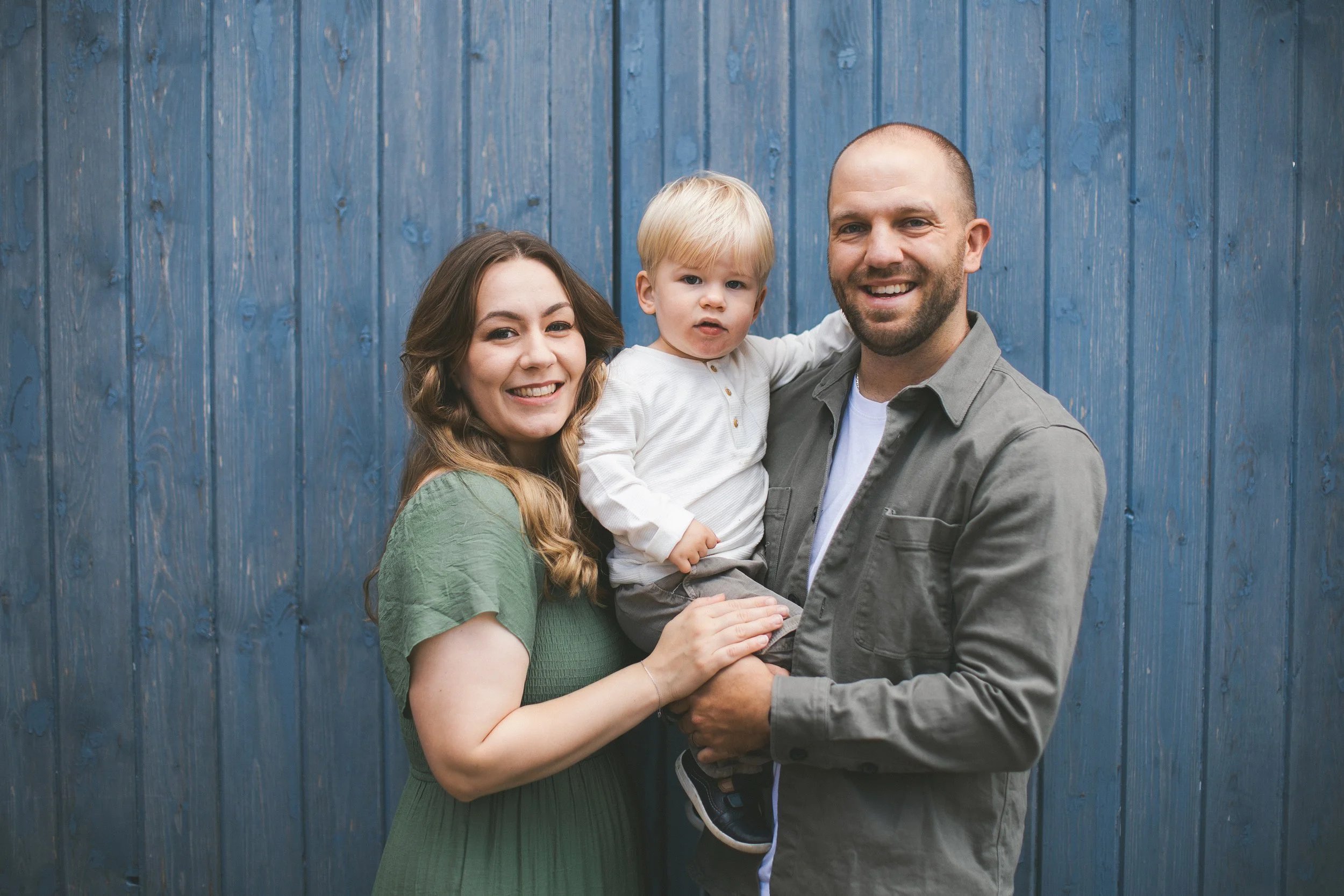 A happy family of three, including a woman with long brown hair, a man with a beard, and a young blonde boy, stands close together in front of a blue wooden fence. The woman and man are smiling, while the boy has a neutral expression.