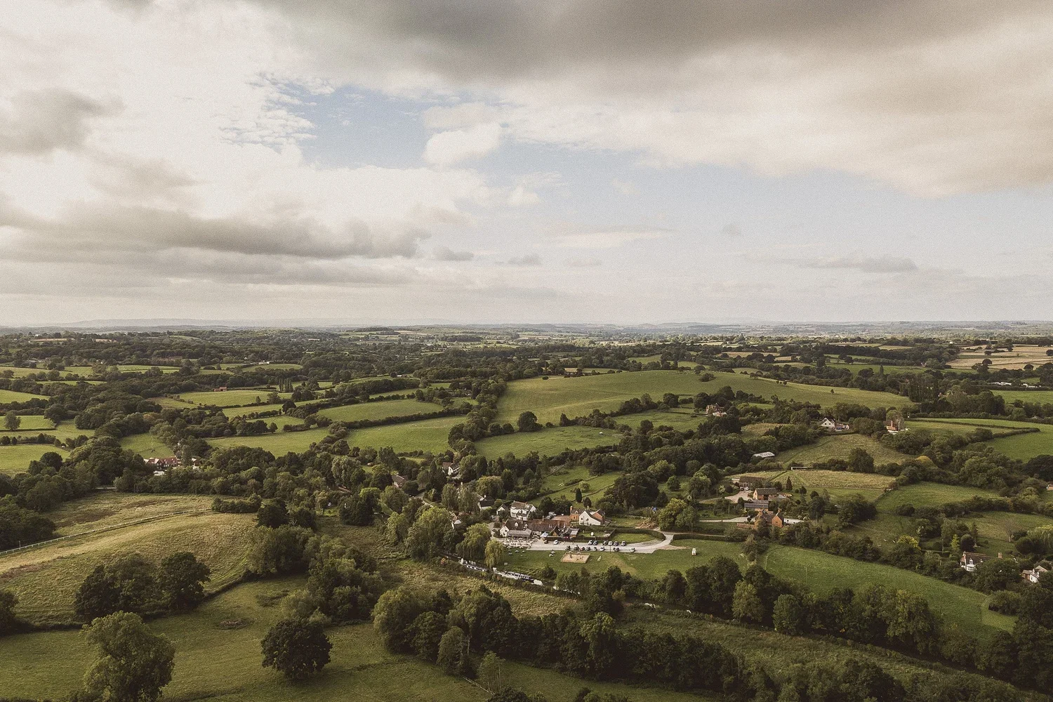 An aerial view of a rural landscape with green fields, trees, scattered houses, and a cloudy sky overhead.