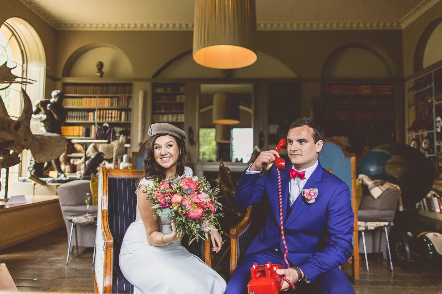 A bride and groom sitting in a decorated room, with the bride holding a bouquet and smiling, and the groom dressed in a blue suit with a pink bow tie, talking on a red vintage telephone.