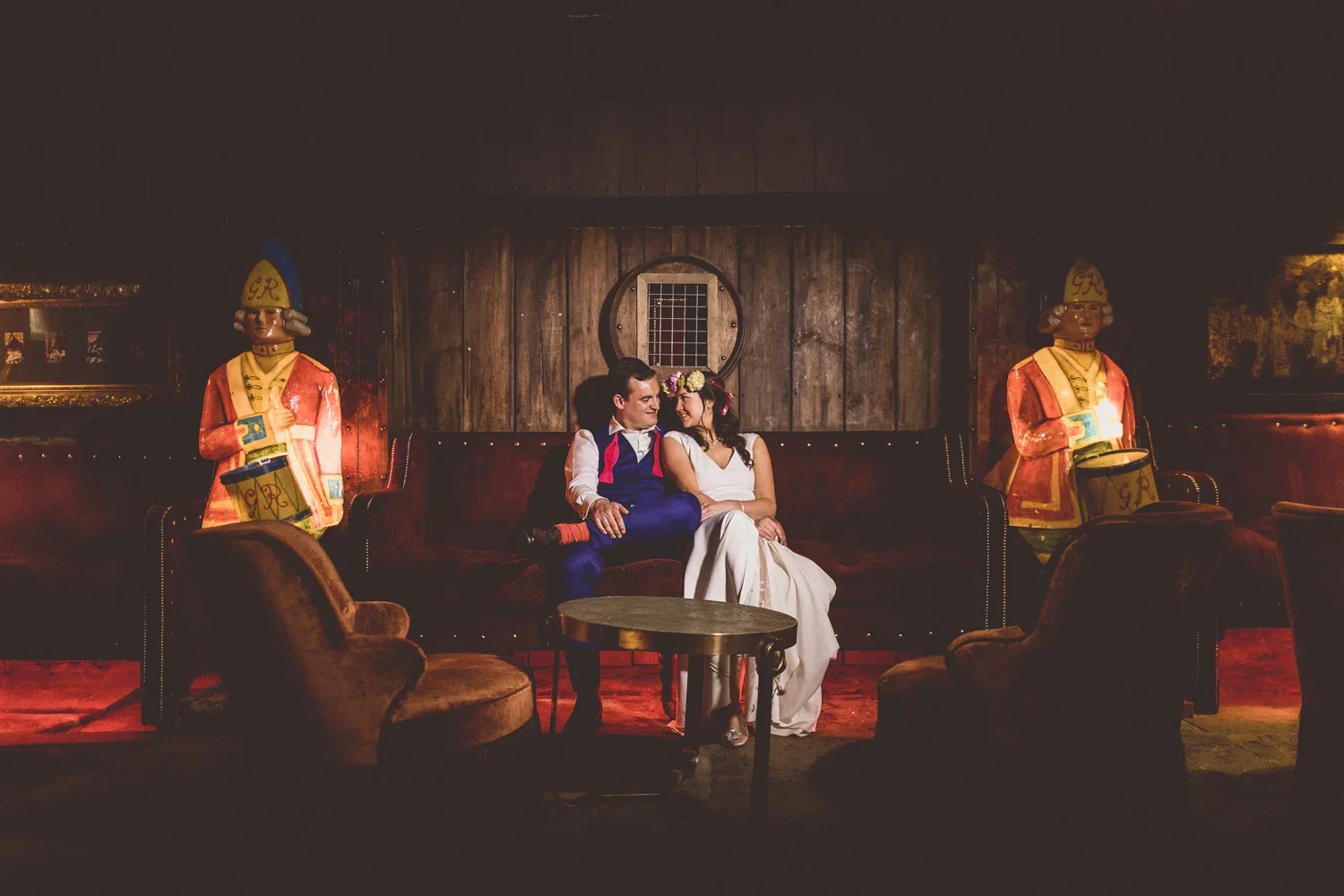 A couple sitting on a red velvet couch in a vintage-style room, smiling at each other, with two large colorful toy soldier statues on either side, and dark wood paneling and paintings in the background.