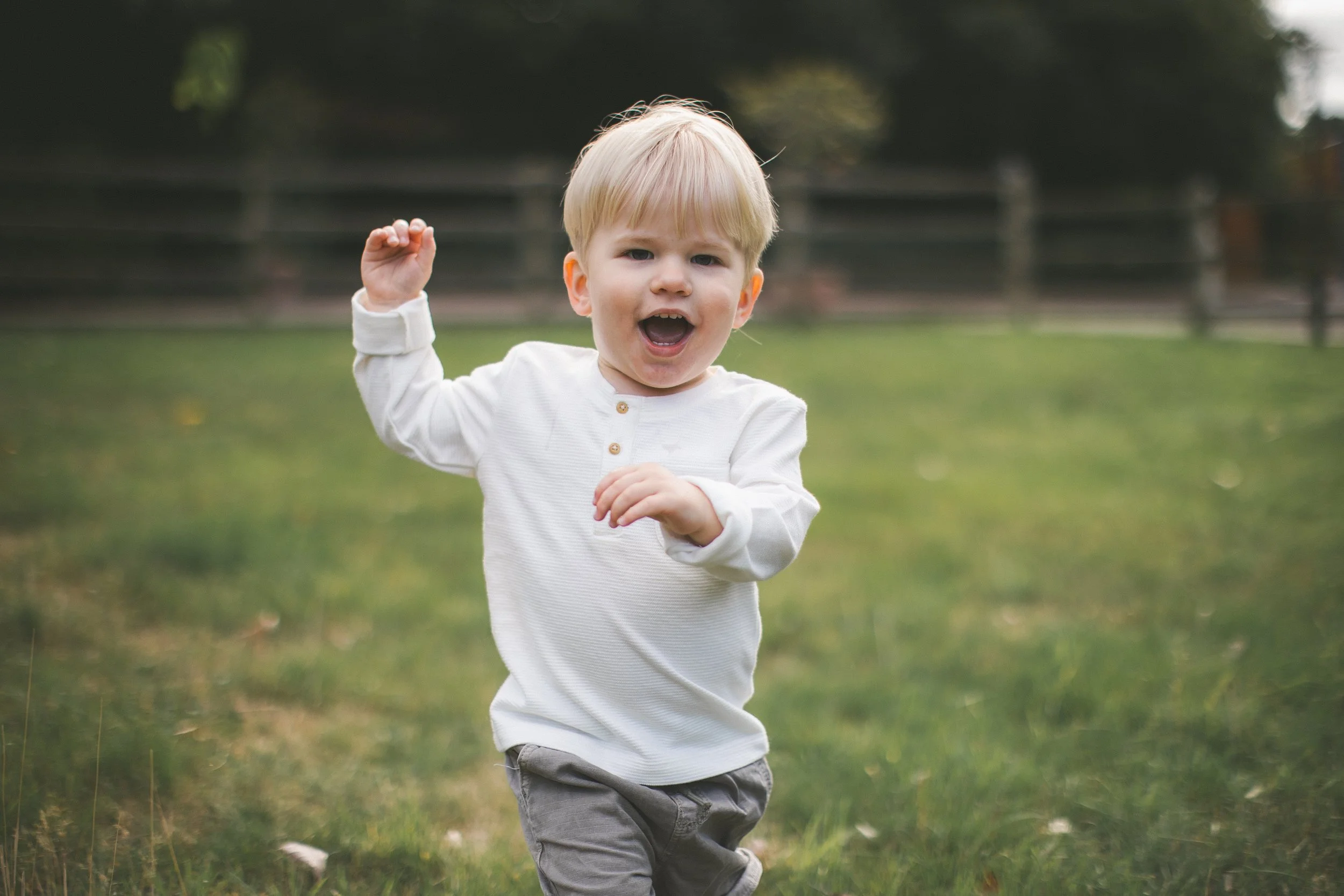 A young boy with blonde hair wearing a white long-sleeve shirt and gray pants is running outside on a grassy field, with a wooden fence and trees in the background.