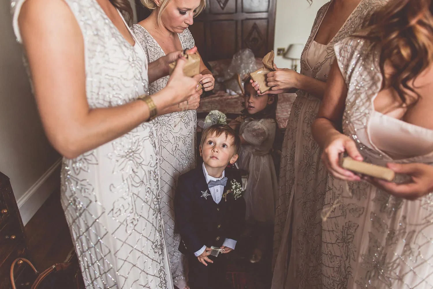 Children at a wedding dress in formal attire, with women in elegant dresses handing out small gift boxes, in a room with a wooden headboard in the background.