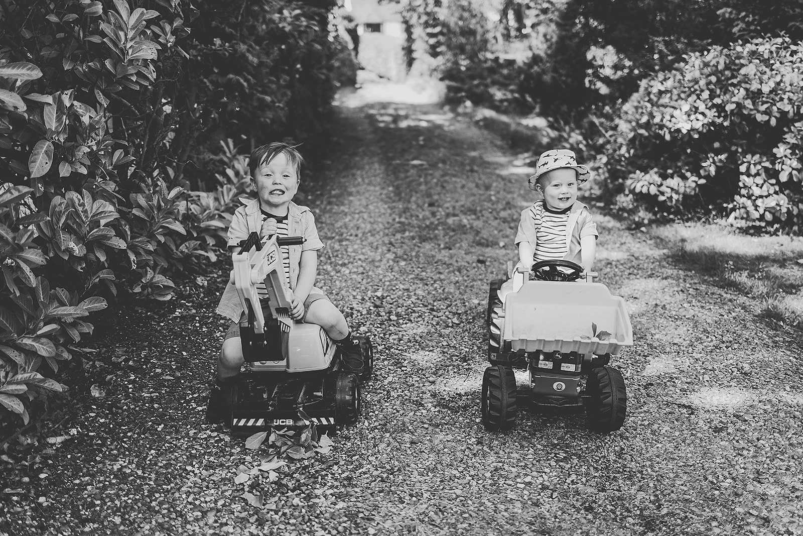 Two young children riding toy construction vehicles on a gravel path surrounded by trees and bushes.