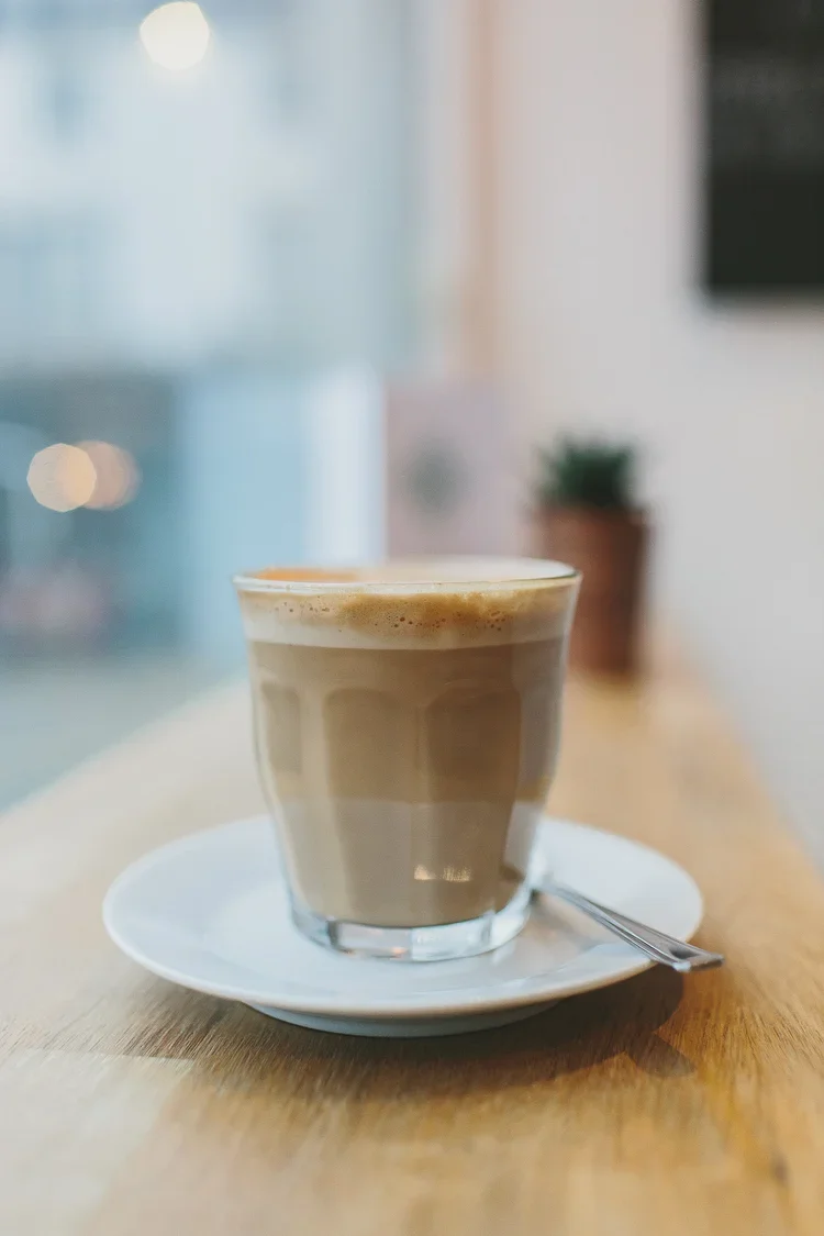 A glass of frothy coffee or latte on a white saucer with a small spoon, placed on a wooden table inside a cafe or coffee shop.
