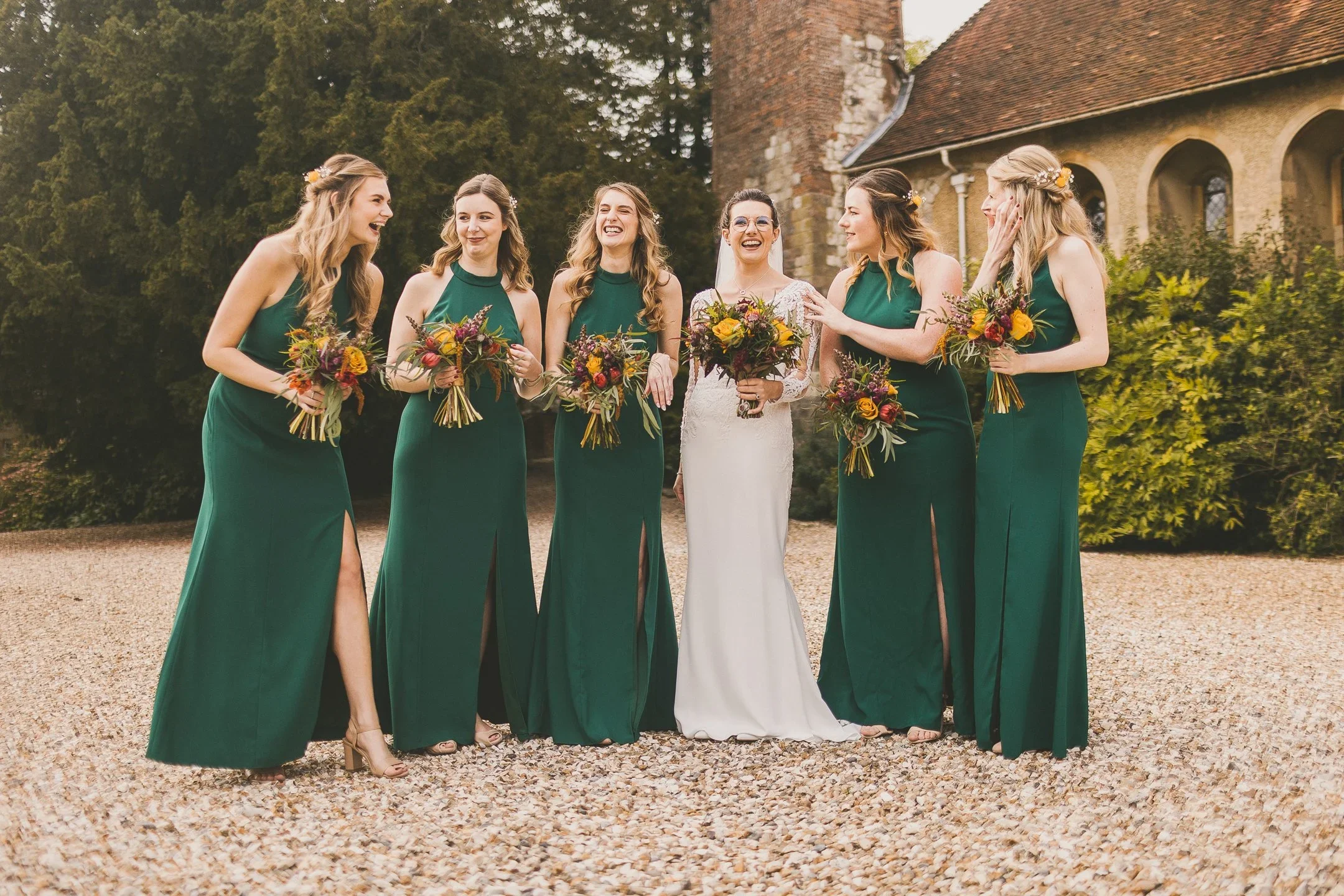 A bride and five bridesmaids standing outdoors, laughing, with the bridesmaids wearing emerald green dresses and holding bouquets, and the bride in a white dress with a veil, against a backdrop of greenery and a stone building.