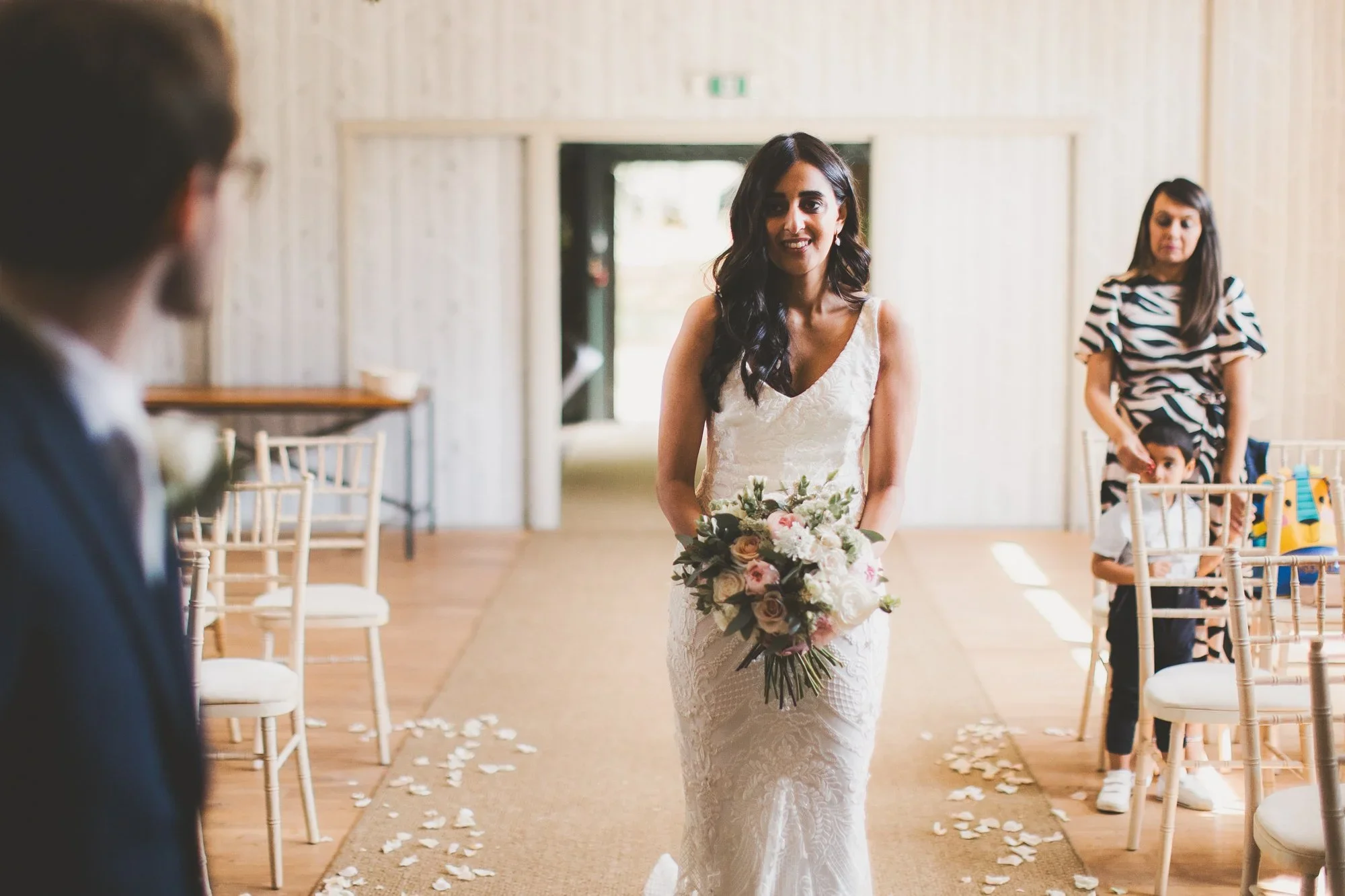 A woman in a white wedding dress holding a bouquet of flowers, walking down an aisle with flower petals scattered on the floor, during a wedding ceremony.