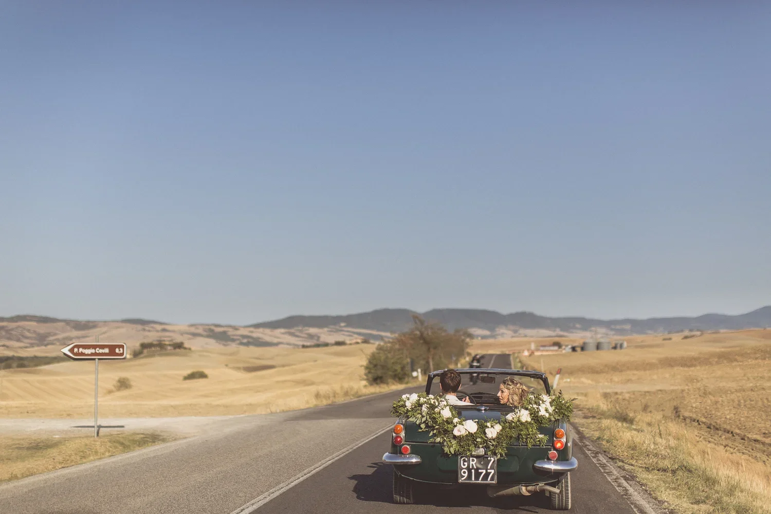 A couple in a vintage convertible decorated with white flowers and greenery, driving on a rural road in a scenic countryside with rolling hills under a clear blue sky.
