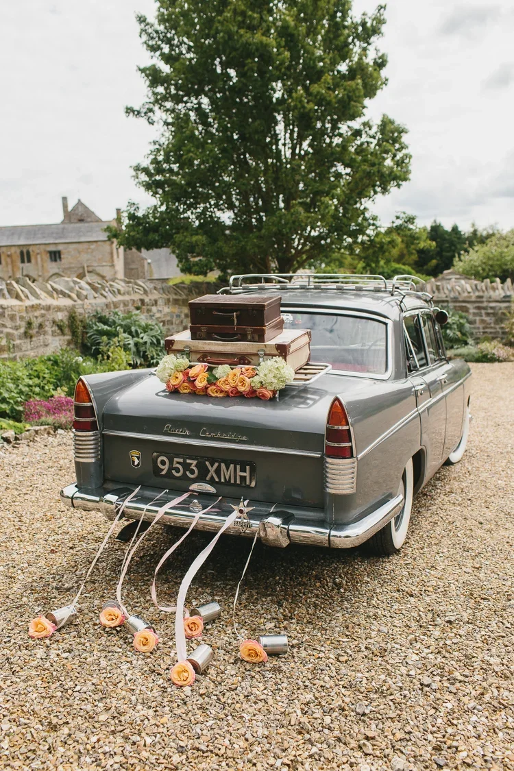 A vintage gray car decorated for a wedding with a flower bouquet, suitcases, and ribbons with roses on the back, parked on a gravel driveway outdoors.