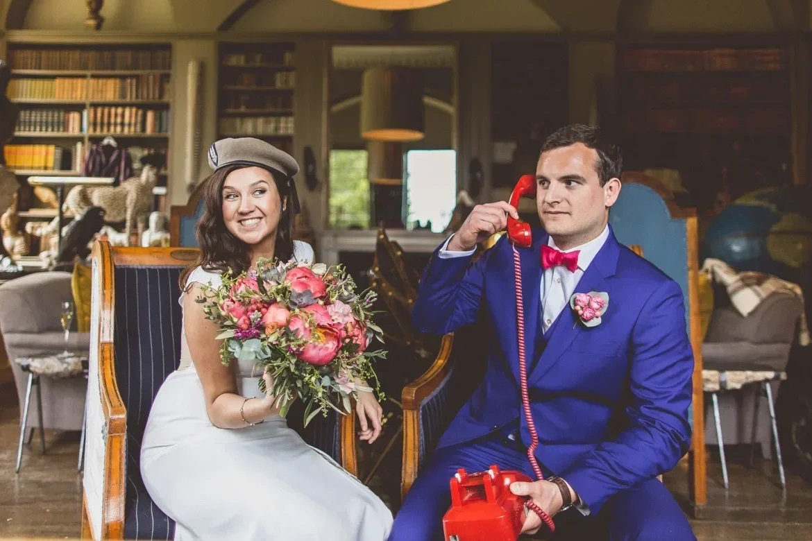 A bride in a white dress and a gray beret holding a bouquet of flowers, sitting next to a groom in a blue suit with a pink bow tie, who is talking on a red vintage rotary phone in a cozy library or living room.