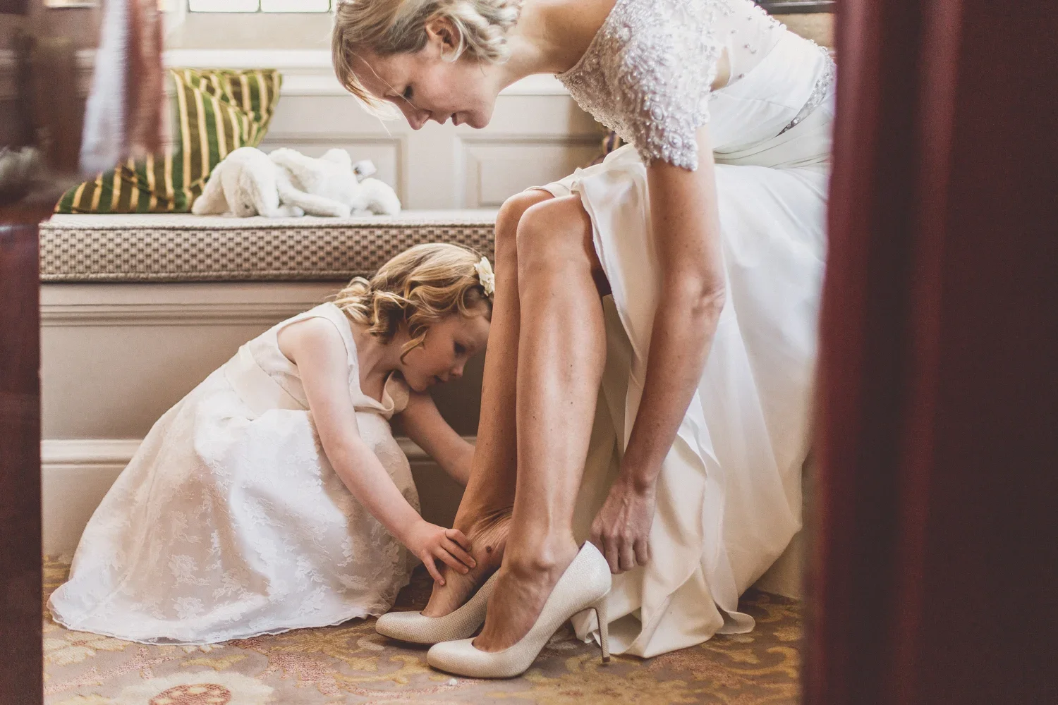 A woman in a wedding dress sitting on a bench, being helped by a young girl in a white dress as they assist her with her high-heeled shoes.
