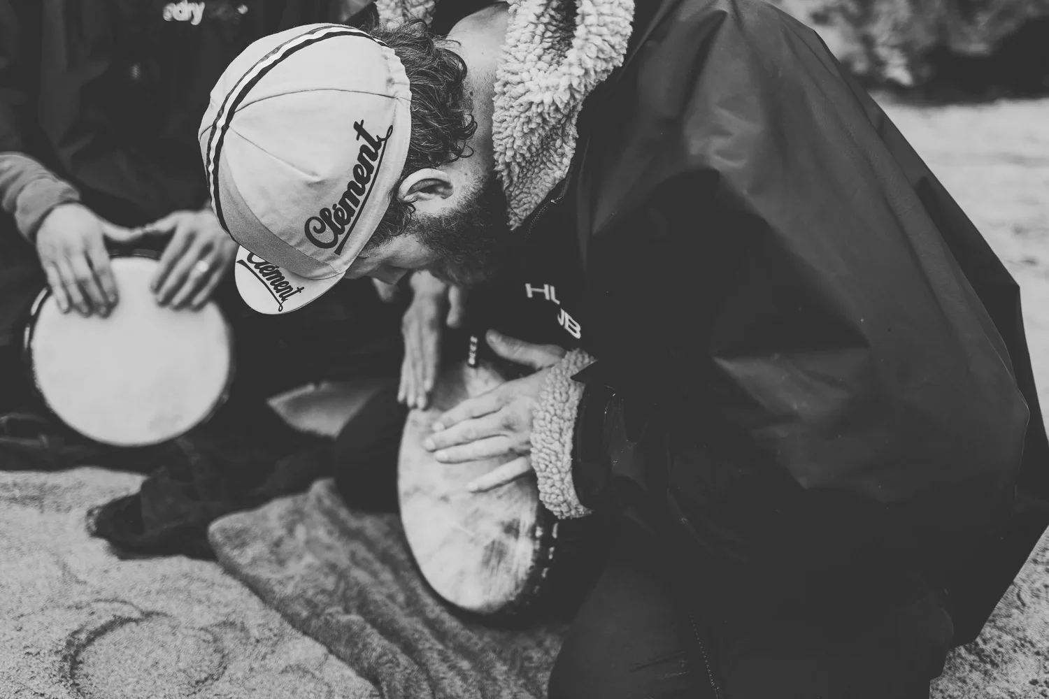 A man with a beard, wearing a baseball cap and a jacket, is performing CPR on a person lying on the ground. The scene appears to be outdoors, and another person is nearby holding a drum.