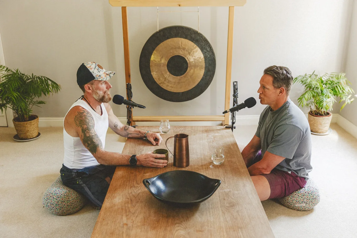 Two men sit across from each other at a wooden table with microphones, glasses of water, and decorative items, engaged in a conversation. They are sitting on cushions on the floor, with plants and a large gong in the background.