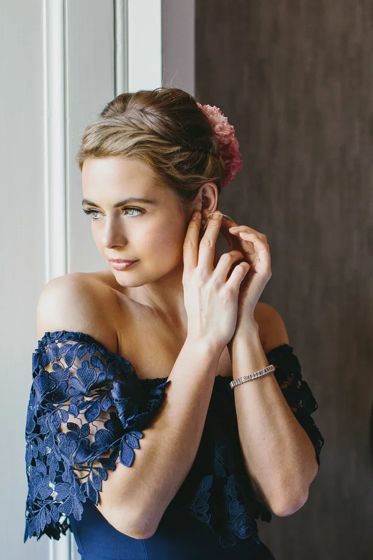 A woman with styled hair adorned with pink flowers, wearing a navy blue lace dress, and putting on earrings by a window.