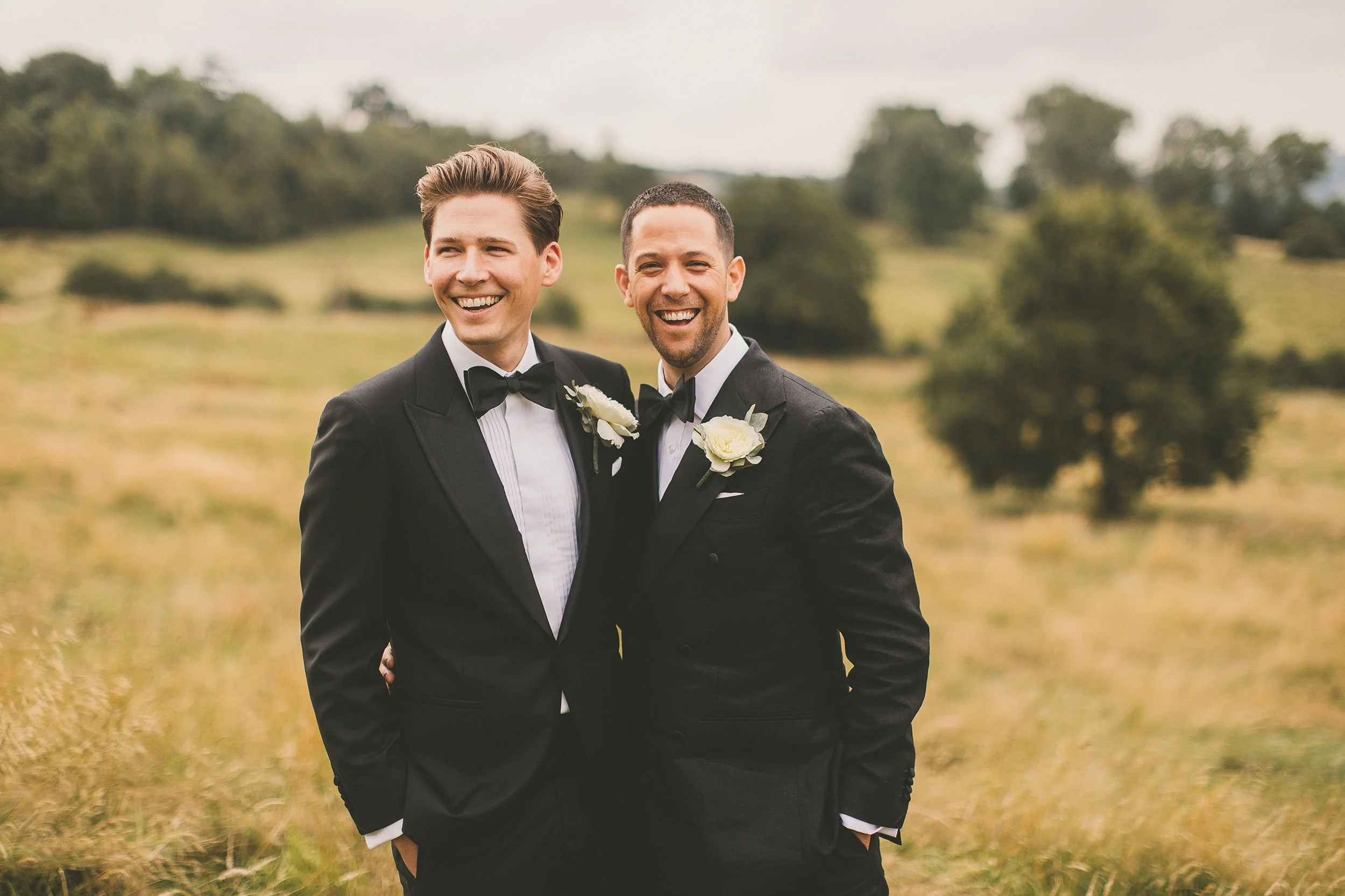 Two men in tuxedos with boutonnières smiling and standing outdoors in a grassy field with trees in the background.