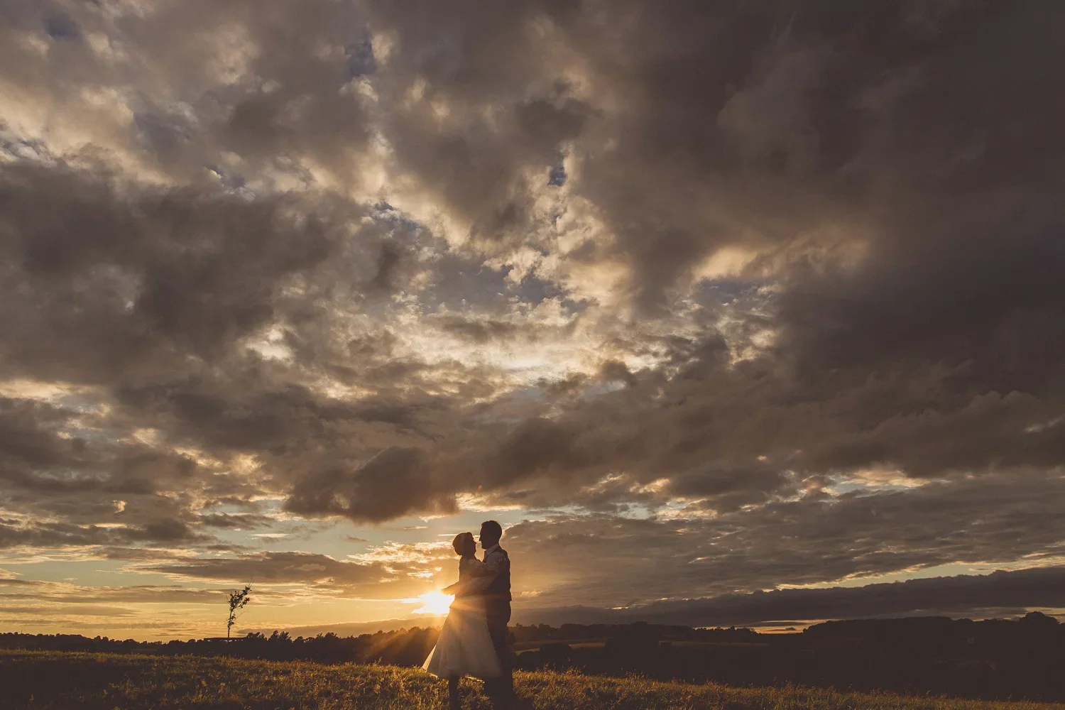 Couple embracing outdoors at sunset, silhouette of man and woman with cloudy sky in background.