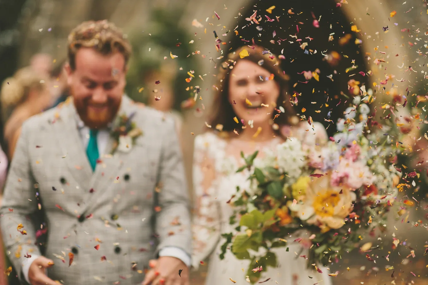 A newlywed couple celebrating their wedding outdoors, with confetti falling around them. The groom is wearing a checked suit and the bride is holding a bouquet of flowers.