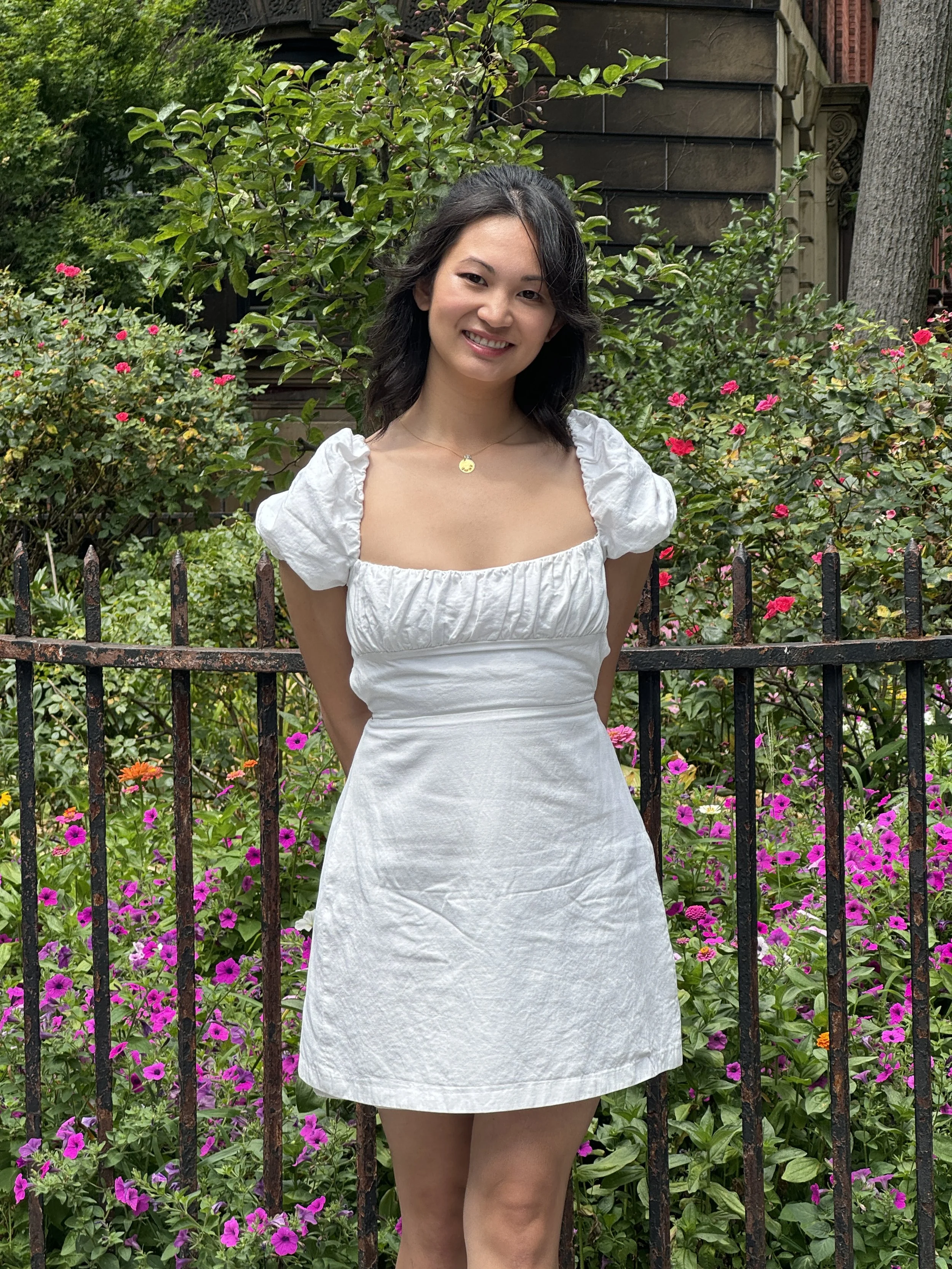 A woman standing outdoors in a garden with pink and purple flowers, wearing a white dress with puffed short sleeves and a gold necklace, smiling at the camera.