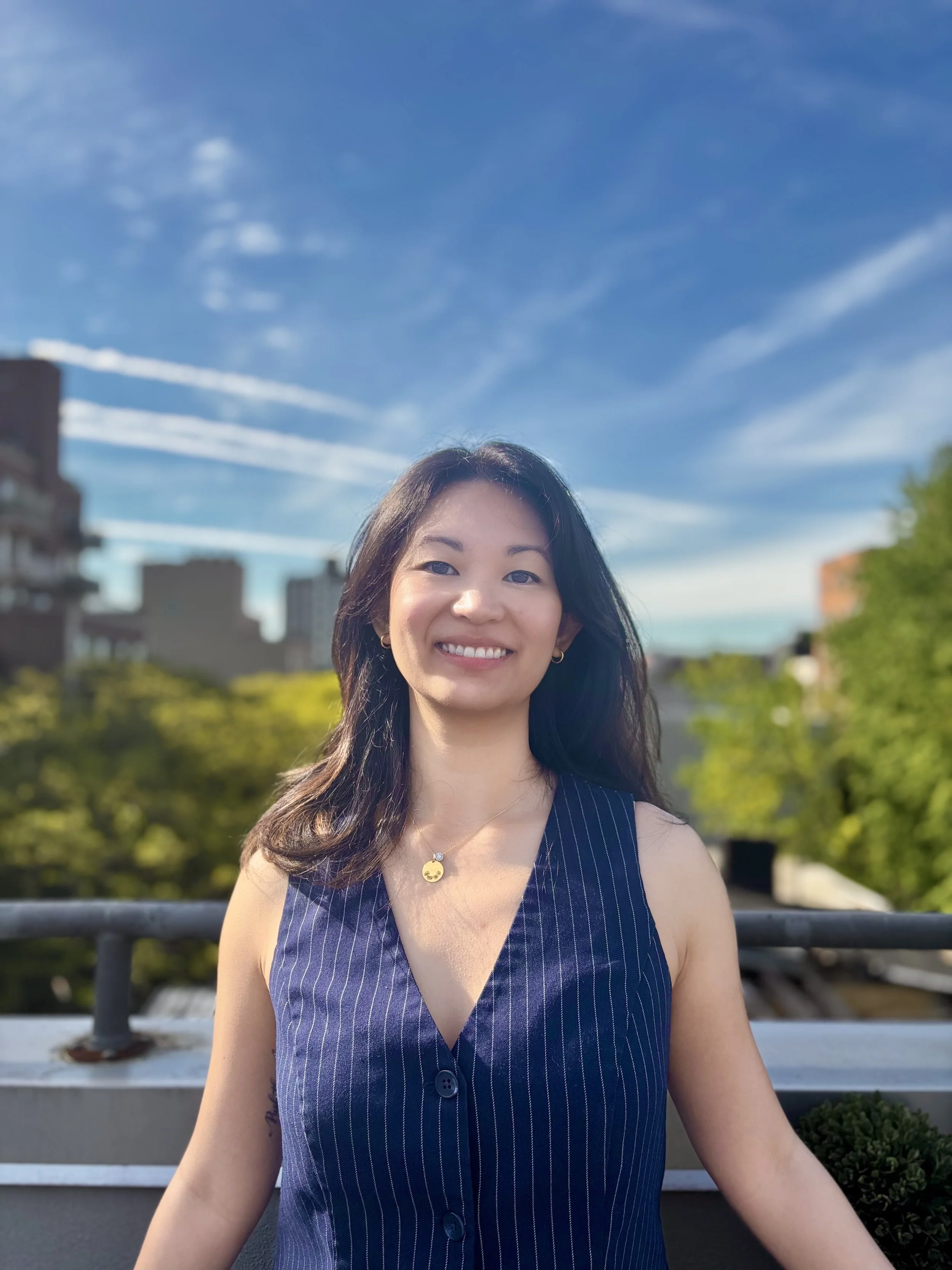 A woman smiling outdoors with a blue sky and wispy clouds, buildings, trees, and a black railing in the background.