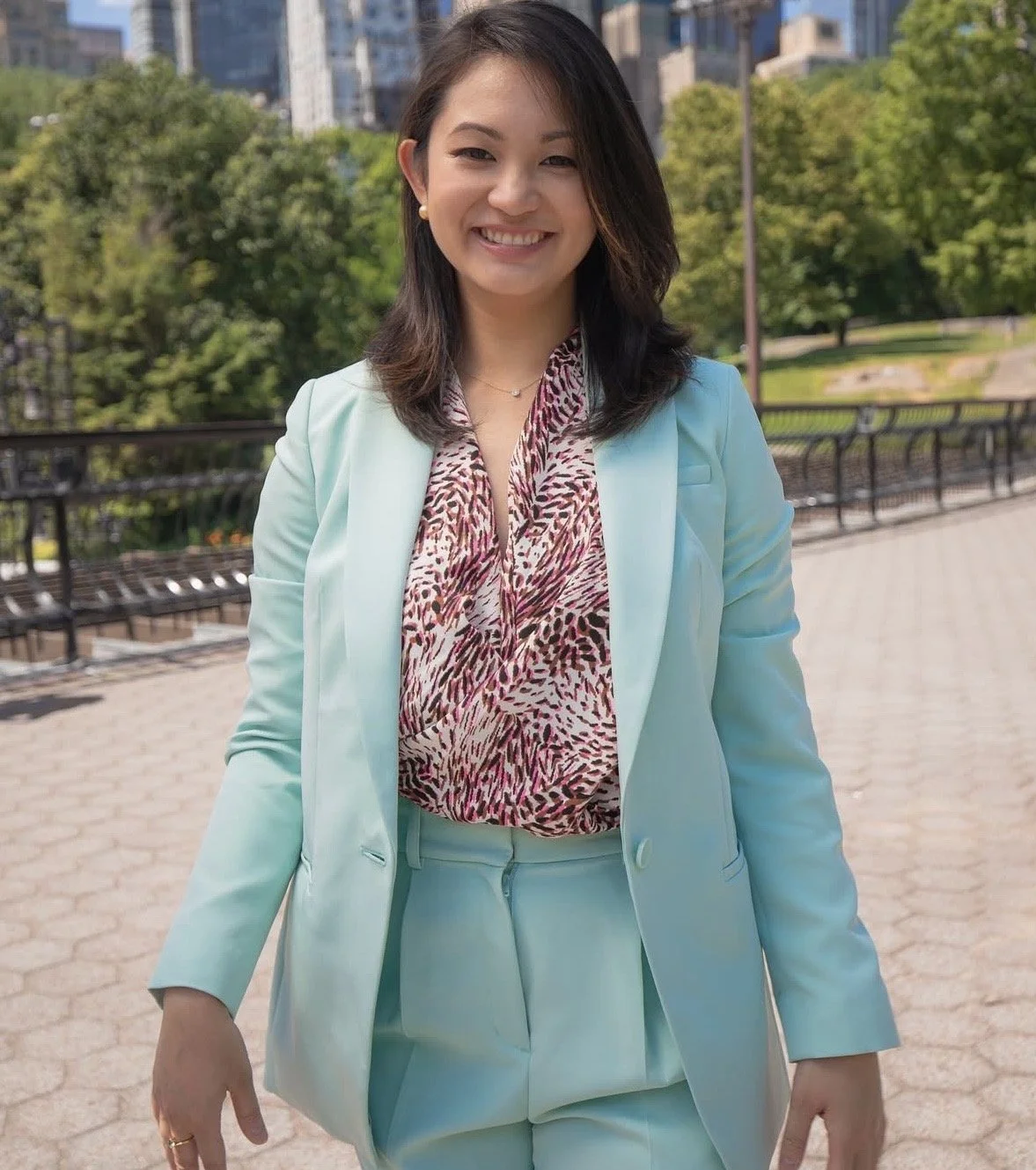 A woman smiling outdoors in a park with trees and city buildings in the background, wearing a light blue suit and patterned blouse.