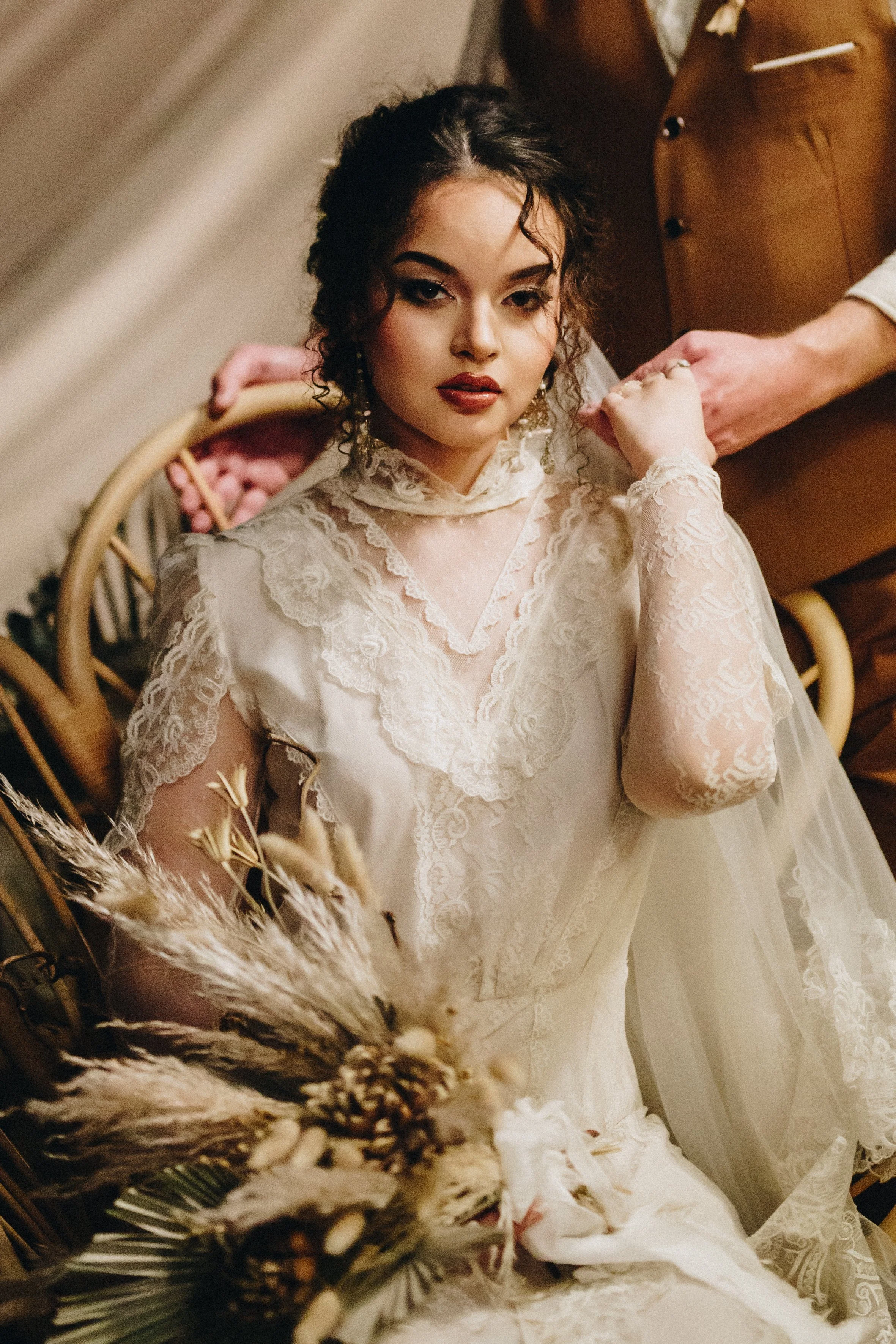 Boho bride sitting in chair with her bouquet on her wedding day with her groom's hand on her shoulder