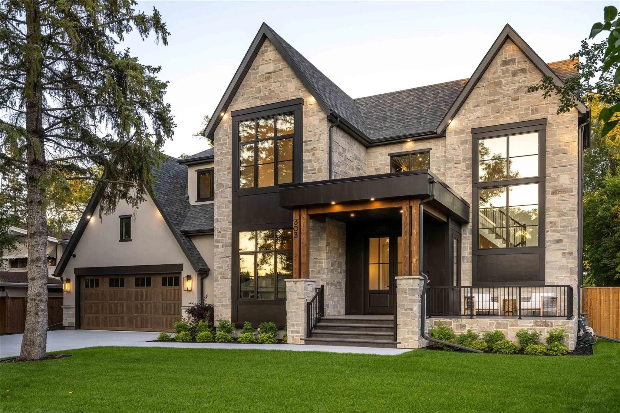 A modern two-story house with stone and black exterior accents, large windows, a front porch, and a well-maintained lawn with shrubs and trees.