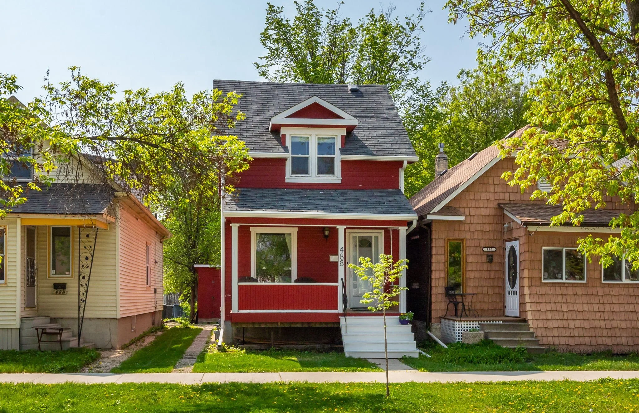 A red wooden house with a small front porch, white stairs, and a dormer window on the roof, situated between two other houses on a tree-lined street.