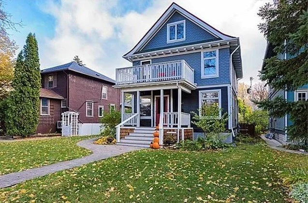 A blue two-story house with a front porch and balcony, surrounded by a lawn with fallen leaves, neighboring houses, and a cloudy sky.
