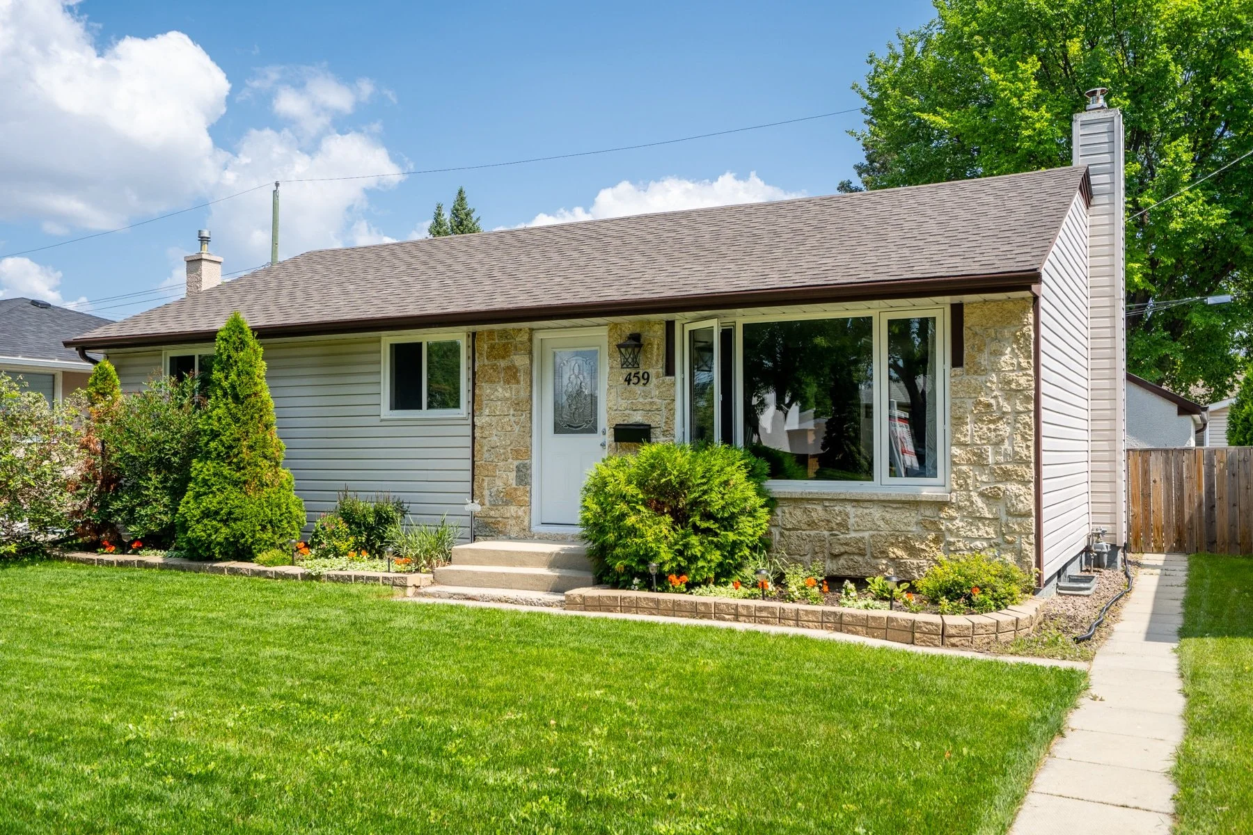 Front view of a single-story house with a brown shingle roof, stone and siding exterior, large front window, and a white front door with house number 459, surrounded by a well-maintained lawn and garden with shrubs and flowers, under a partly cloudy sky.