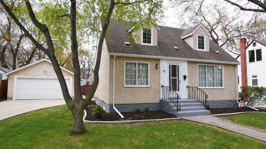 A beige two-story house with a front porch, black iron railings, and steps leading to the front door. There are large windows, and a detached garage is visible to the left. The yard has green grass, a tree, and some shrubbery, with a brick walkway leading to the front steps.