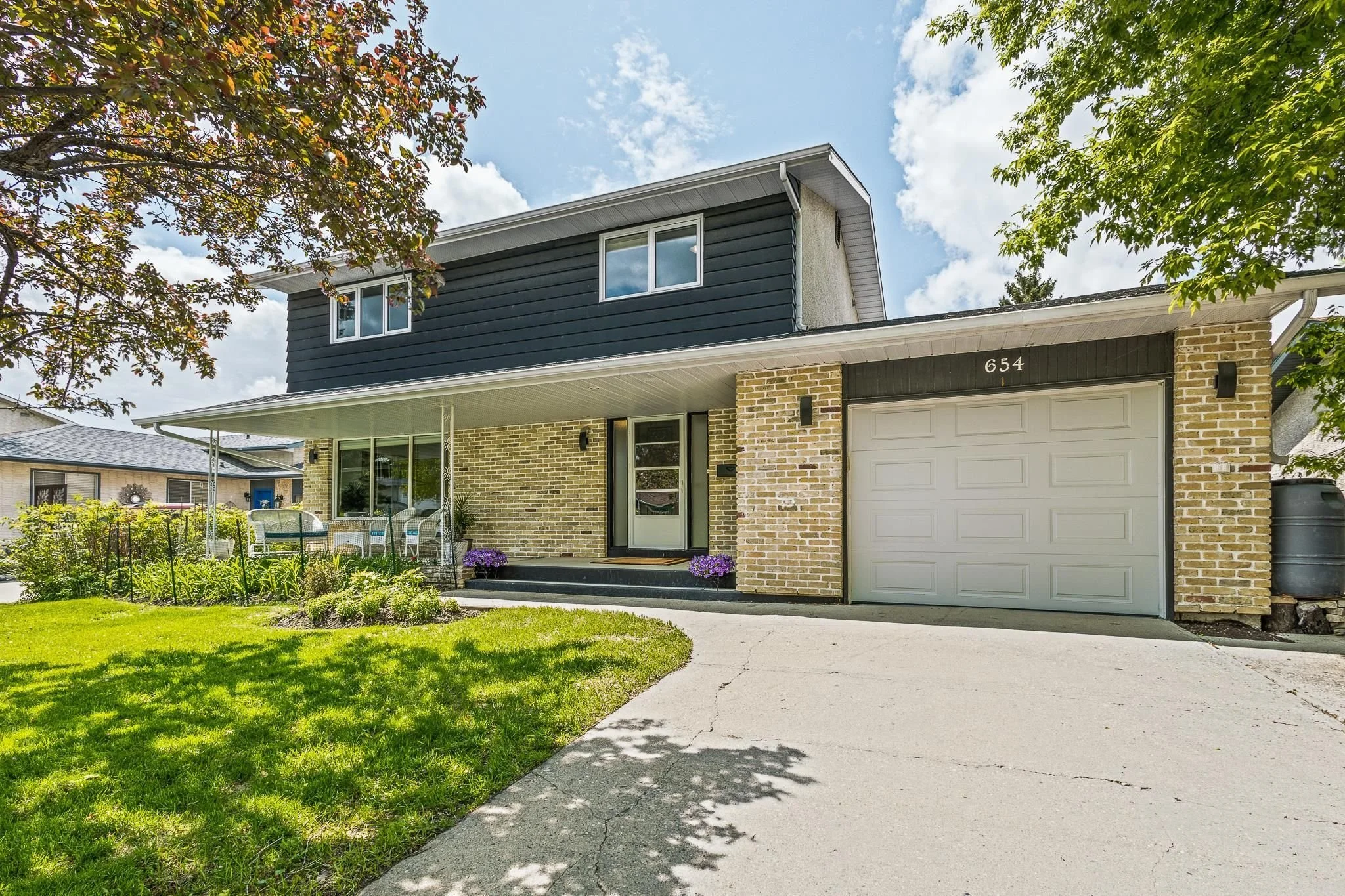 A two-story house with a brick front and dark blue siding on the upper level, a white garage door, and a small porch with outdoor seating. There are trees and a green lawn in front, with a concrete driveway leading to the garage.