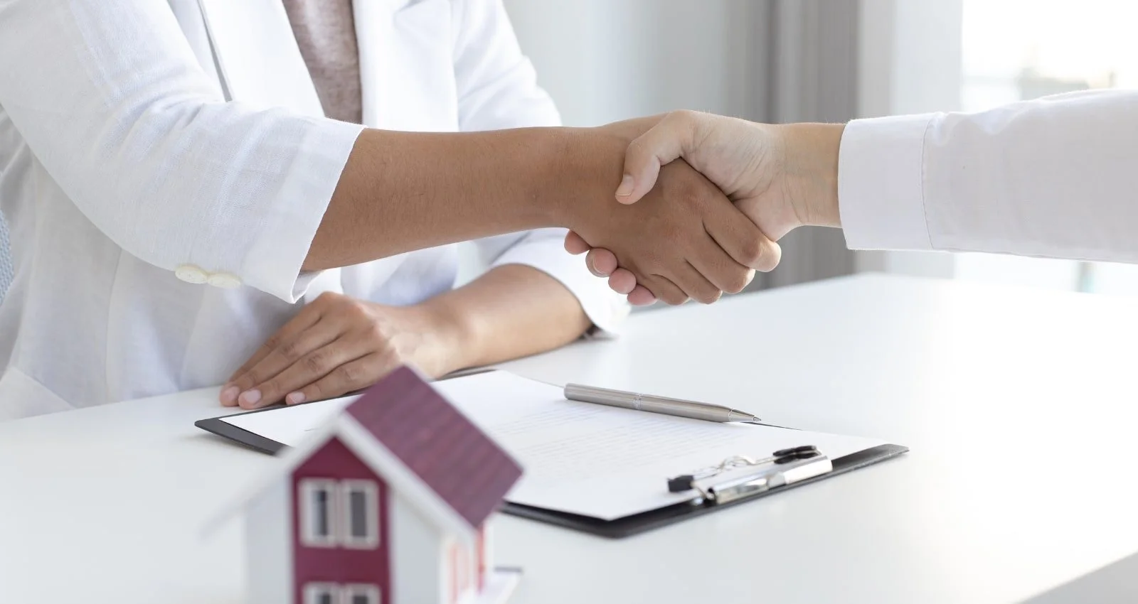 Two people shaking hands over a desk with a clipboard, a pen, and a small house model.