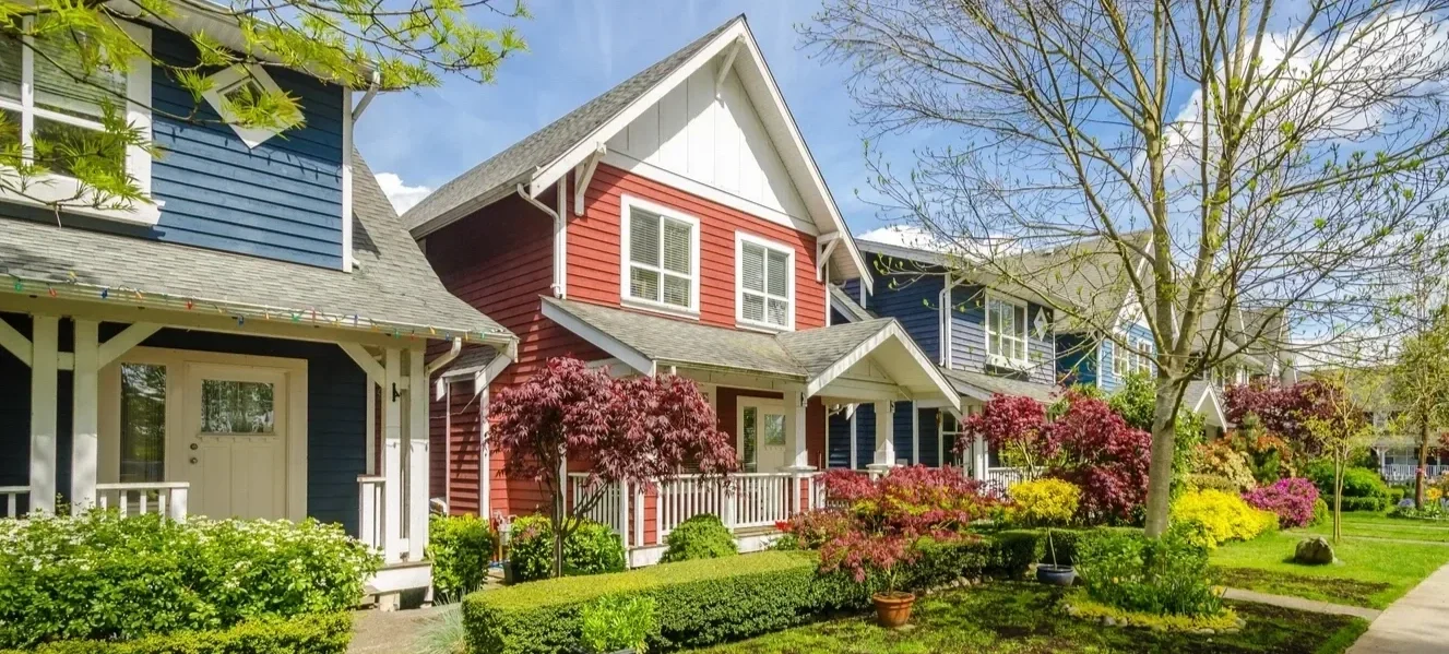 Colorful row of houses with lush gardens, trees, shrubs, and a sidewalk on a sunny day.