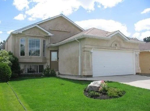 Front view of a two-story suburban house with beige exterior and a white garage door, surrounded by a green lawn and a flower bed.